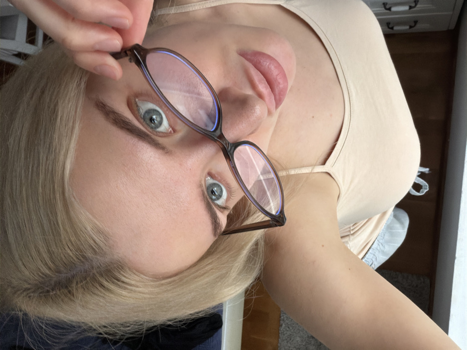 A close-up, high-angle portrait depicts a fair-skinned female individual with light blonde hair and prominent light blue eyes. She looks directly into the camera. Her right hand, with visible fingernails, holds a pair of brown-framed eyeglasses by the temple arm, positioned across her face, with the lenses partially obscuring her left eye and resting near her nose and upper lip. The lenses exhibit multiple light reflections. Her lips are full. She wears a light beige or nude-colored camisole with thin shoulder straps. The background is blurred, showing indistinct interior elements including a dark horizontal surface and a white, possibly paneled, object. The lighting appears to be natural and originates from the front-left. This scene is located in the city of Budva, Montenegro.