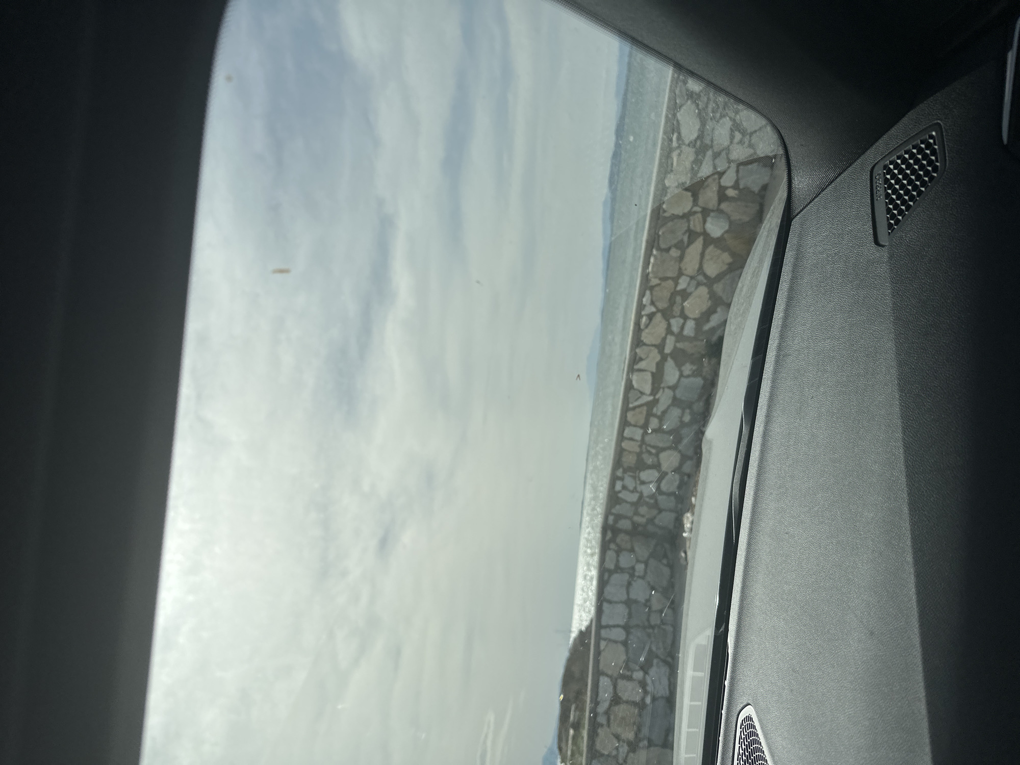 The image captures a serene coastal or lakeside scene, viewed from inside a vehicle, likely in Fanari, Greece. The photograph appears to be rotated ninety degrees clockwise, showing the horizon vertically.
In the foreground, the dark gray, textured interior paneling of the car is visible on the right and bottom edges. A speaker grill with a honeycomb pattern is prominently featured on the upper right side of this panel. Faintly visible above this grill are the letters "AIRBAG". Another speaker grill is partially visible at the bottom right. The window frame of the vehicle outlines the view to the outside.
Through the window, a body of water, presumed to be the sea given the location context, occupies the left and central portions of the frame. Its surface reflects light, creating a bright, shimmering effect. Along the edge of this water, there is a distinct rocky or paved path, possibly a seawall or promenade, constructed from irregularly shaped, light-colored stones. In the distant background, faint landforms or mountains rise beneath a muted, overcast or partly cloudy sky.
The scene is set during the daytime, with soft, diffused natural light illuminating the landscape. The weather appears to be calm, with subtle ripples on the water's surface. No specific human activity is depicted, emphasizing a moment of tranquil observation from within the car. The only visible text is "AIRBAG" on the car's interior.