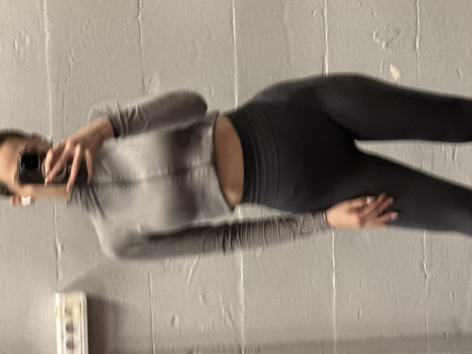 A person stands in front of a grey wall, holding a cell phone to take a selfie. They wear dark leggings and a grey zip-up jacket. The location cannot be determined from the image, and Sofia, Bulgaria cannot be confirmed.