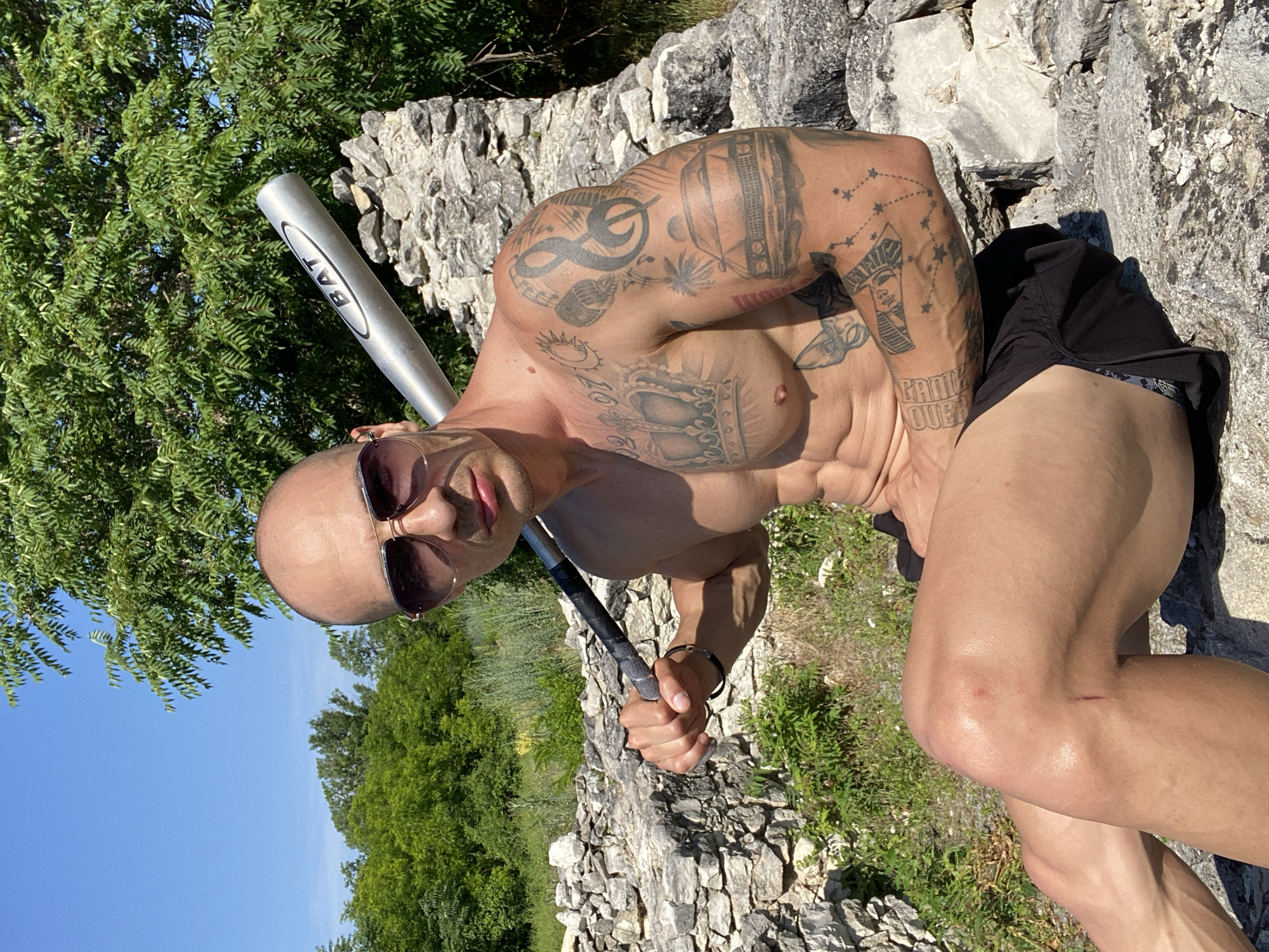 A shirtless, muscular bald man with sunglasses is sitting on rocks outdoors. He is holding a baseball bat over his shoulder. The man has extensive tattoos on his arms and chest. He is wearing black shorts. The background consists of lush green trees and blue sky. The scene appears to be in a rural or historical outdoor setting, possibly an archaeological site or ruins, with stone walls visible. The lighting suggests it is a sunny day. While the location is stated to be in Pleven, Bulgaria, there are no specific landmarks within the image to confirm this.