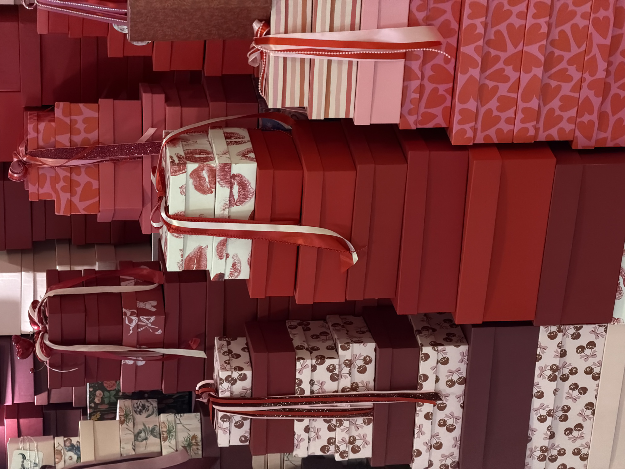 The image shows a wall adorned with numerous stacked gift boxes of varying sizes and designs, primarily in shades of red, pink, and white. Some boxes are decorated with patterns such as hearts, kisses, stripes, and cherries. Many are tied with ribbons. The exact location is not discernable, as the image lacks visual cues that would definitively identify Moscow, Russia.