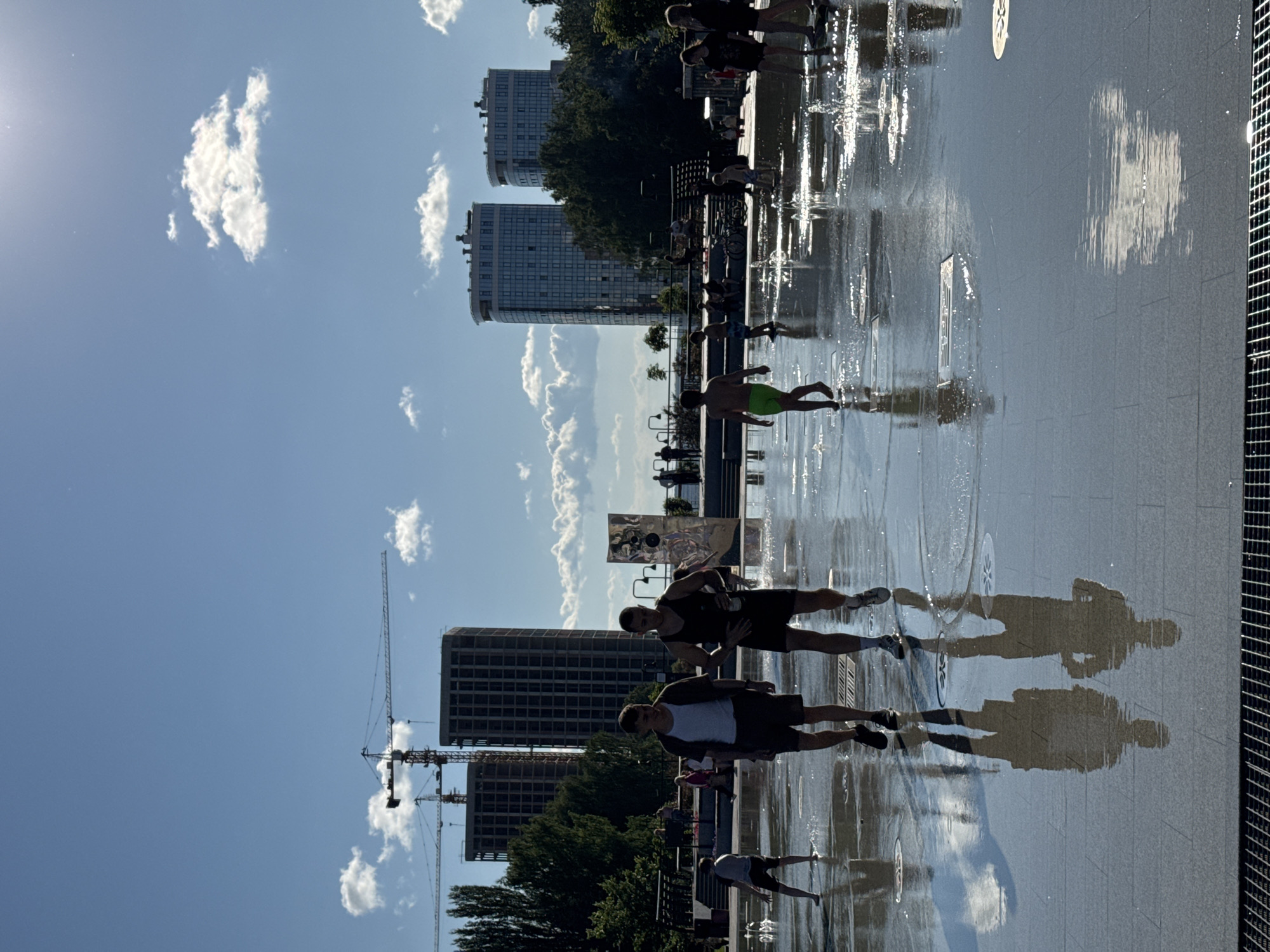 The image shows a group of people, including children and adults, enjoying themselves in a splash pad or public fountain area. The ground is wet, with water spraying up in various locations. Several individuals are walking through the water, and some appear to be playing. In the background, there are modern buildings, some of which appear to be under construction with cranes visible. Trees and the sky with clouds are also present. The overall scene depicts a sunny day in an urban outdoor recreational space. The location is the splash pad in the "Gala" park on the embankment in Izhevsk, Russia.