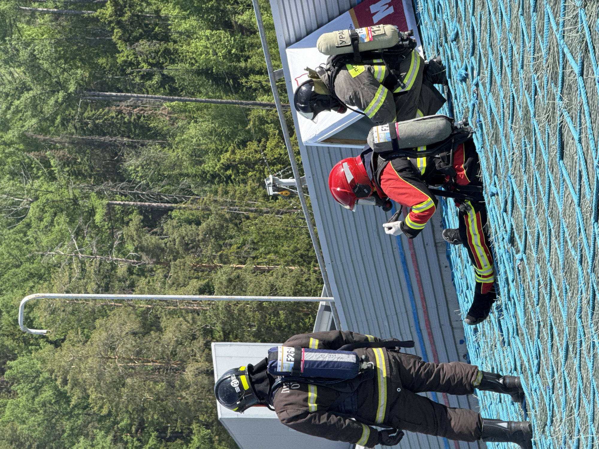The image captures three firefighters on a blue safety net during what appears to be a training exercise. They are wearing helmets, protective gear and breathing apparatus. The background includes a metal structure, a lush green forest, and a glimpse of what may be buildings or infrastructure. The location is Unknown.