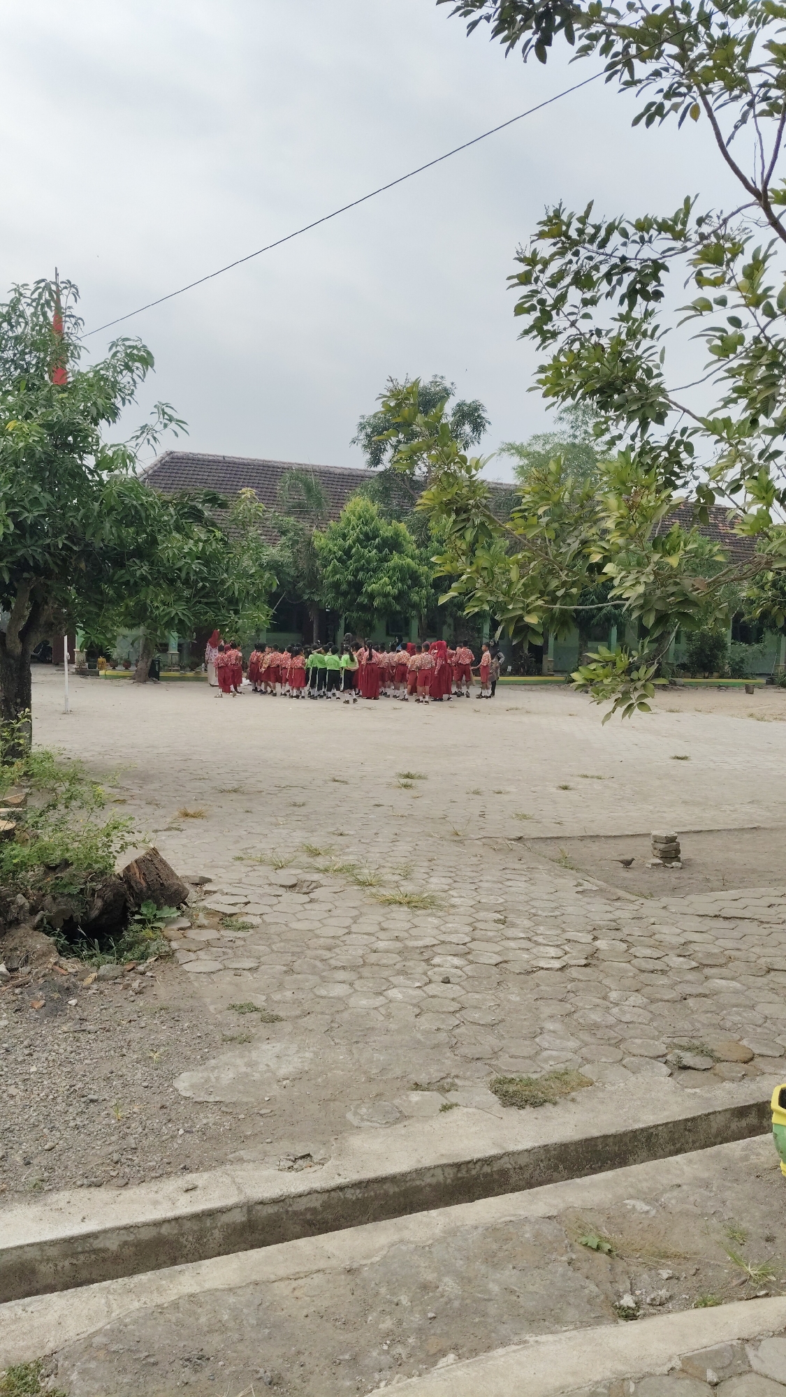 In Banjarsari, Indonesia, a group of schoolchildren in red and green uniforms stand in formation on a paved courtyard in front of a low-rise building with a tiled roof. Trees with green foliage are visible around the courtyard and the building. The sky is overcast. The foreground shows a concrete drainage ditch.