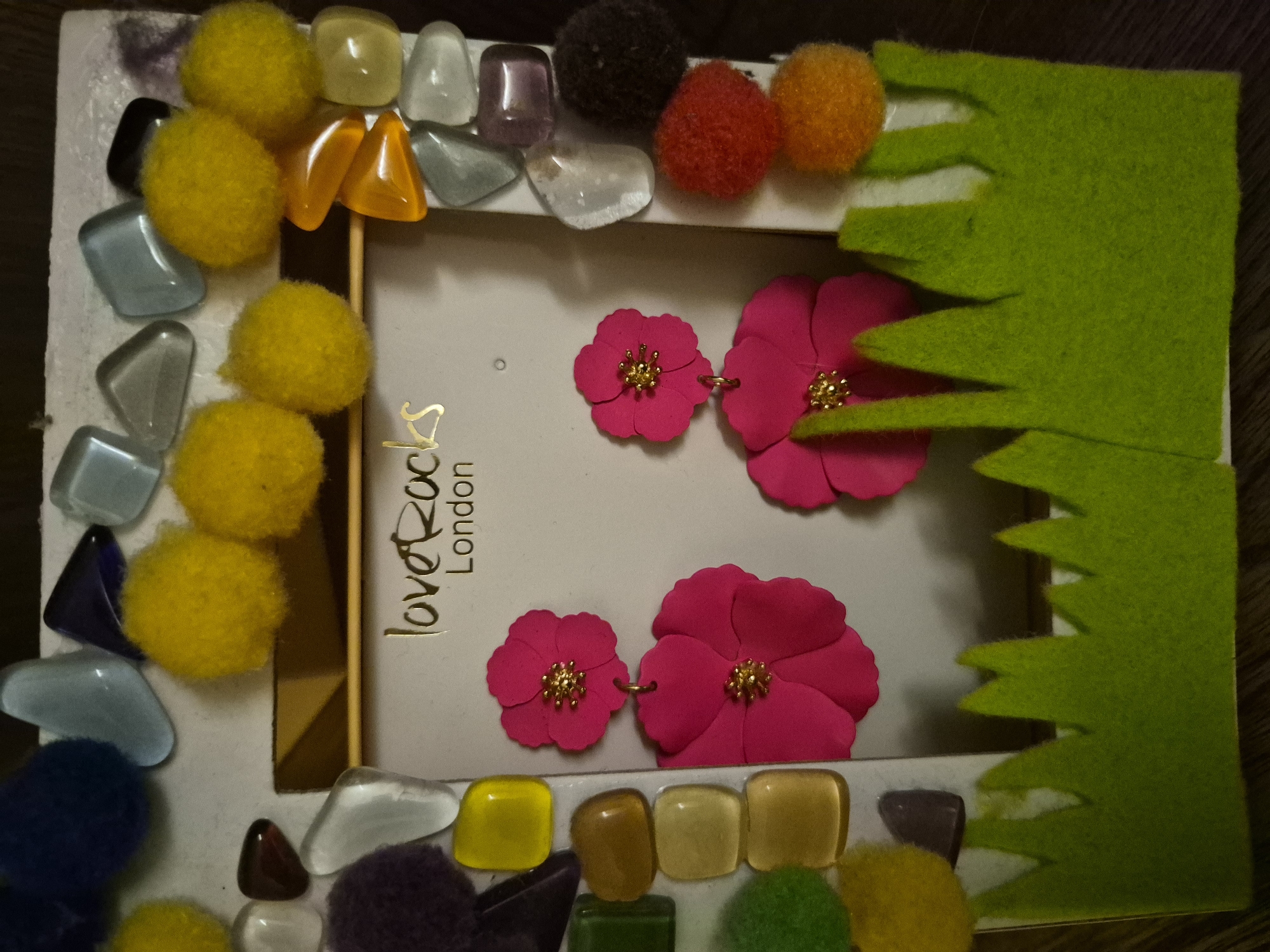 The image shows a close-up, eye-level, indoor shot of a decorative picture frame. The frame is adorned with various colorful objects, including smooth, colorful stones and fluffy pom-poms in shades of yellow, blue, purple, and red. A pair of bright pink flower-shaped earrings with gold centers hangs from the center of the frame. The background of the frame displays the "Love Rocks London" logo. The bottom of the frame is decorated with a piece of green felt cut to resemble grass. The overall scene is static and appears to be a still life arrangement. The location is not specified.