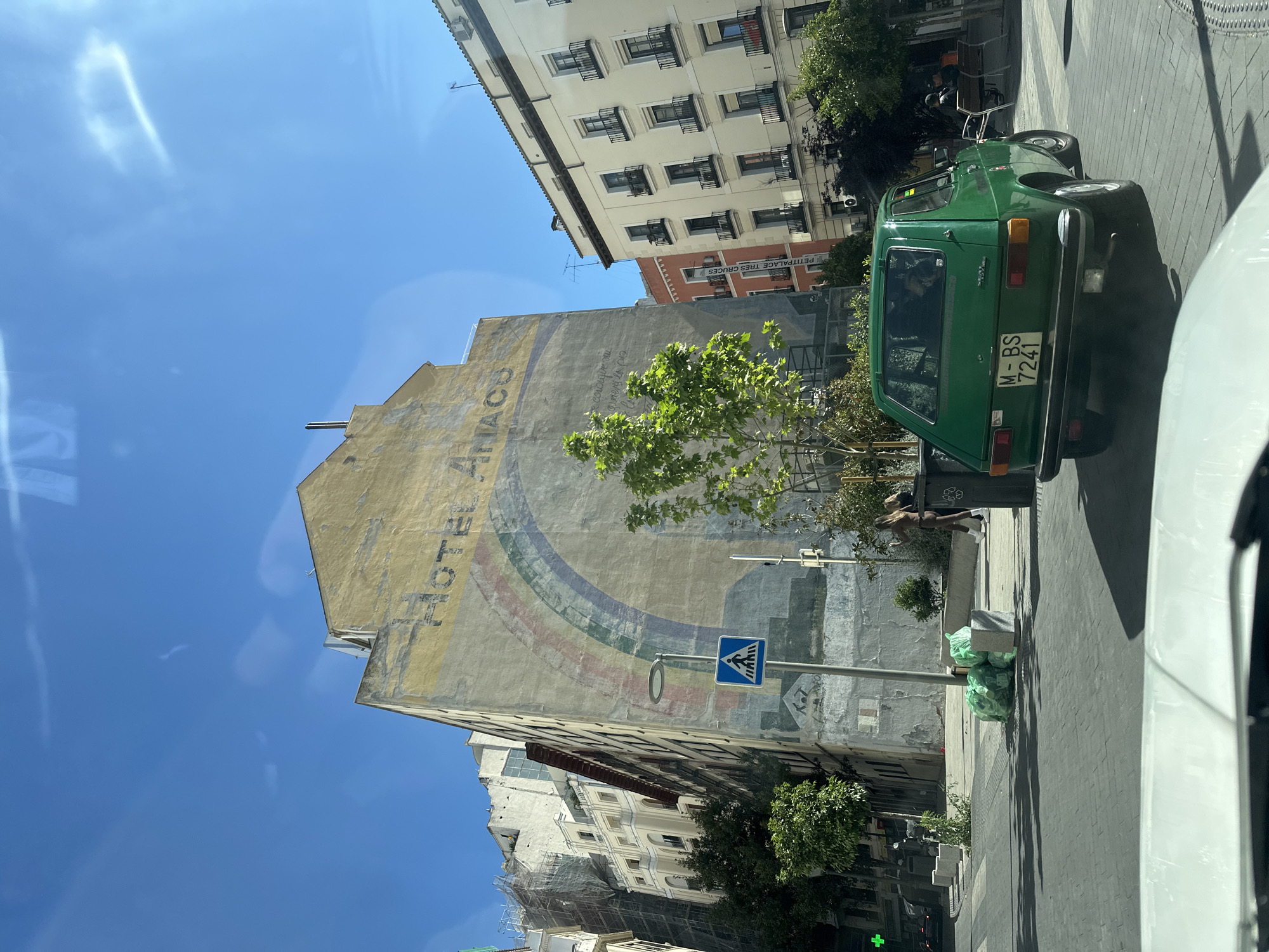 The image shows a street scene in Madrid, Spain. A faded advertisement for "HOTEL ANACO" is painted on the side of a tall building with a rainbow design. A green vintage car with the license plate "M-BS 7241" is parked on the street. A pedestrian crossing sign is visible. There are green garbage bags on the sidewalk. Buildings with multiple windows and balconies are in the background. The sky is clear and blue.