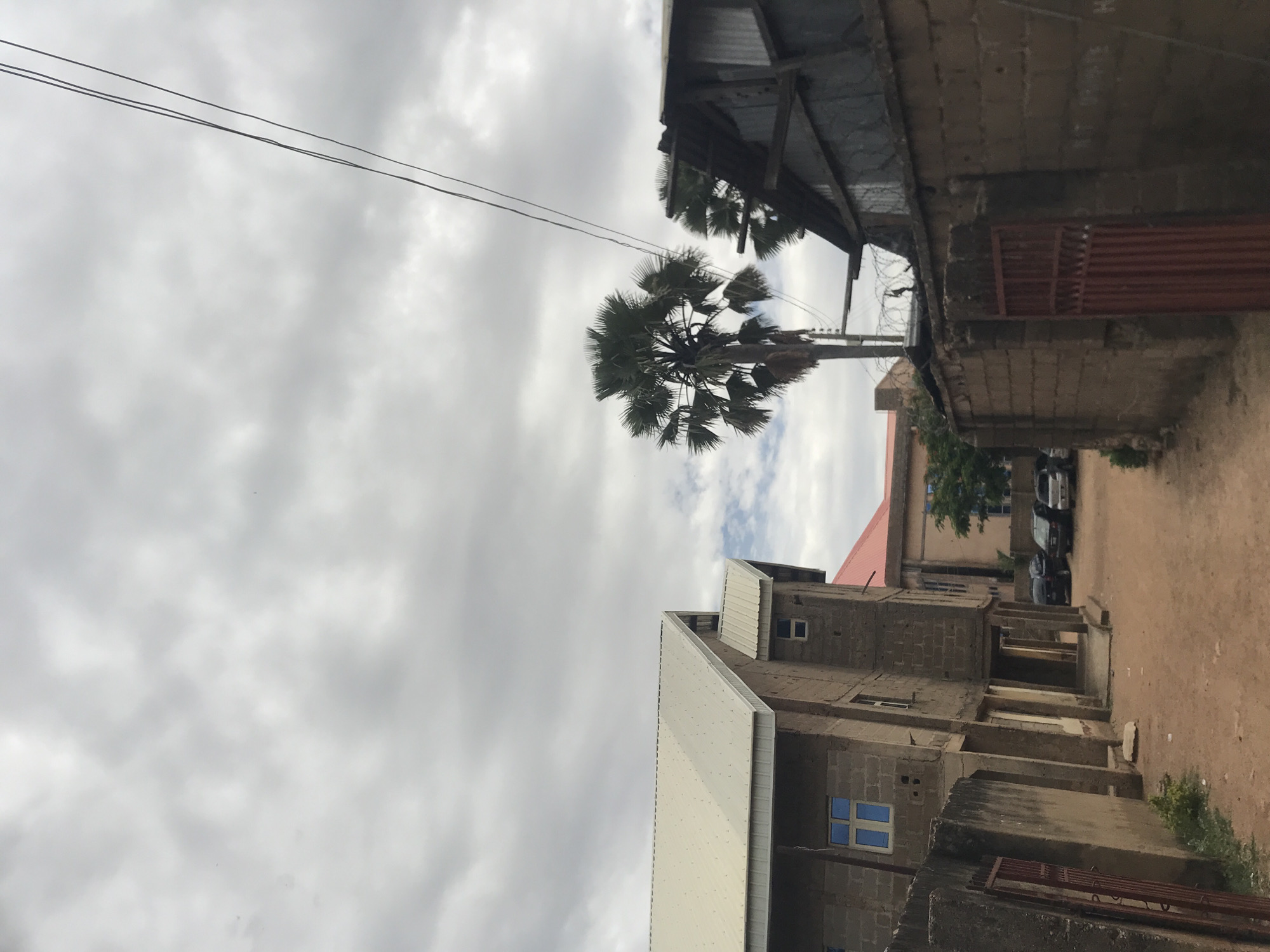 The image depicts a semi-urban scene in Rafin Par, Nigeria, under an overcast sky filled with dense grey clouds. The foreground features a dirt road or path leading into the distance. On the left, a multi-story building constructed from concrete blocks is visible, appearing to be either under construction or recently completed, with a prominent light-colored corrugated metal roof and blue-tinted windows. An older, similarly constructed section is attached below.
On the right, a weathered wall built from concrete blocks runs parallel to the path. This wall is topped with barbed wire or razor wire, and behind it, a structure with a corrugated metal roof and sides is partially visible. A red, vertical-slat metal gate is set into this wall. In the midground, a tall palm tree with fan-shaped fronds stands prominently, with another smaller palm tree partially obscured behind the corrugated structure on the right. Utility poles and power lines traverse the upper portion of the scene. Further in the background, a building with a red roof and several parked cars are discernible. No individuals are present in the frame.