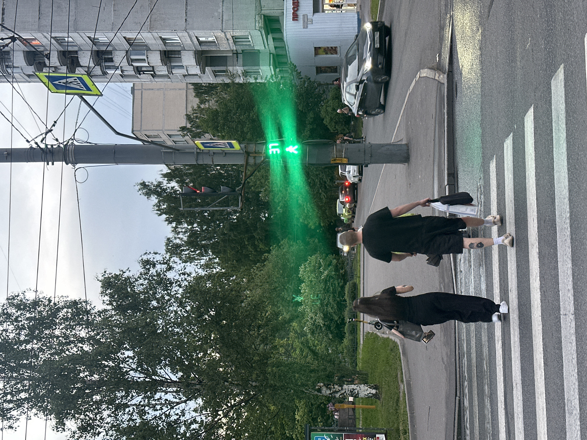 The image depicts an urban street scene at dusk or early evening in the city of Unknown, Unknown. Two pedestrians, a female and a male, are captured mid-stride, walking away from the camera on a white-striped zebra crossing. The female, on the left, wears a black top and pants with white sneakers, carrying a dark tote bag and holding a smartphone. The male, on the right, wears a black t-shirt, black shorts, and light sandals with white socks, carrying a white plastic bag and another dark bag in his right hand; a dark tattoo is visible on his left calf.
A prominent grey utility pole stands centrally, adorned with multiple wires and two yellow-and-blue pedestrian crossing signs. A pedestrian traffic light attached to this pole displays a bright green walking figure and the number "37," casting a strong green glow across the scene. To the left, a vehicle traffic light shows red.
On the right side of the road, a dark-colored sedan with illuminated headlights is in motion, driving from right to left. Further along the street to the left, several vehicles are stopped or parked, including a light-green car, a dark-red sedan, and a silver car.
The right side of the frame features a multi-story, grey concrete apartment building with enclosed balconies; an orange light is visible in one upper-floor window. Adjacent to this building is a single-story commercial structure with a sign partially visible in Cyrillic script, reading "ПРОДУ" (indicating "products") and "БОГАТЫРСК..." with the number "5". Several indistinct figures are visible near this shop entrance.
The left side of the scene is dominated by dense green foliage from numerous trees, including a distinctive birch tree with white bark. Bushes line the sidewalk and road divider. An electric scooter is parked on the sidewalk near the birch tree. Overhead power lines crisscross the top portion of the frame against a light grey, overcast sky. The asphalt road appears damp in various spots.