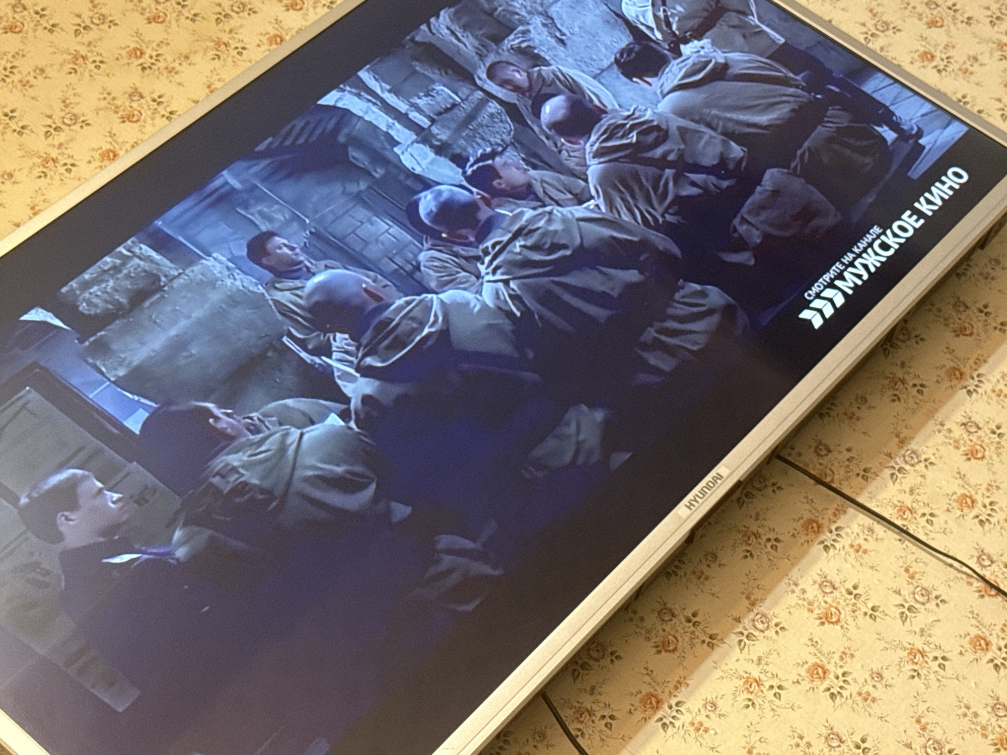 The image shows a Hyundai television displaying a scene of people in what appears to be a military setting, possibly a movie scene. The people are predominantly wearing olive-green military-style clothing. They are crowded together, some sitting or kneeling. The environment looks like a subterranean concrete structure. The text on the screen says "СМОТРИТЕ НА КАНАЛЕ МУЖСКОЕ КИНО", which translates to "Watch on the men's cinema channel." The TV is resting on a floral-patterned surface. The image was taken in Mytishchi, Russia.