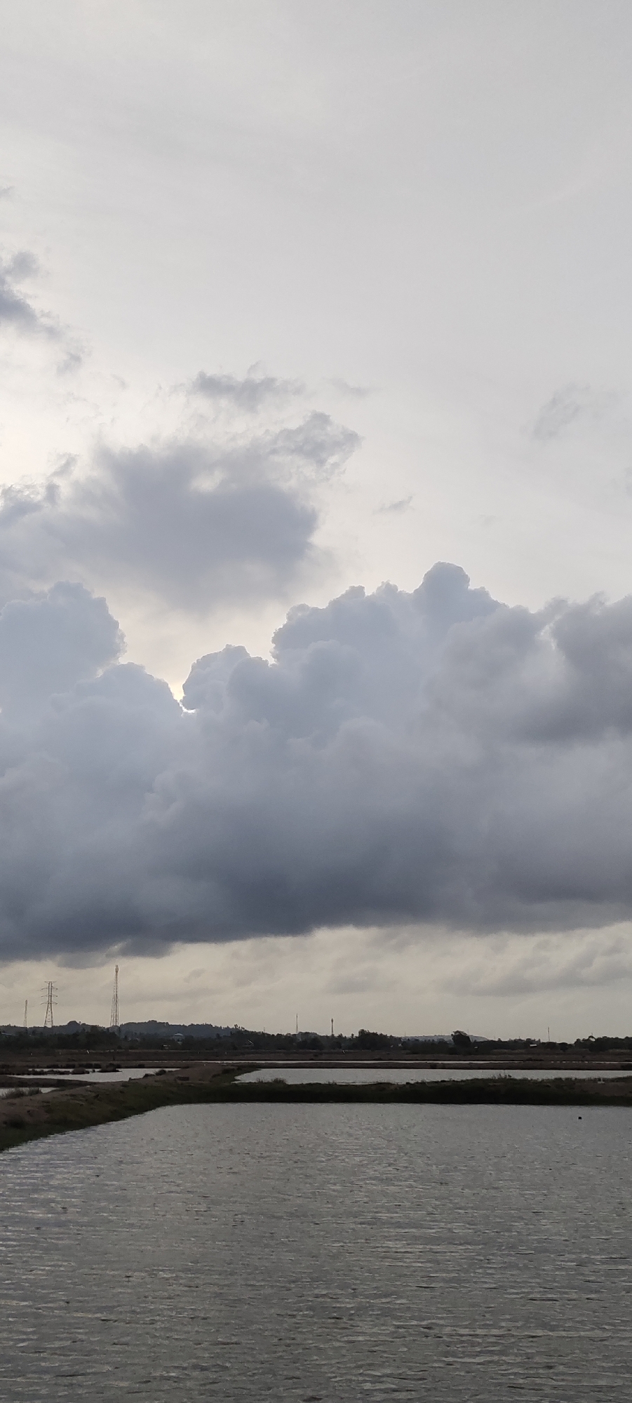 The image depicts a landscape in Lhokseumawe, Indonesia under an overcast sky dominated by heavy, grey cumulonimbus clouds. The lower portion of the scene features a body of water, likely a series of ponds or wetlands, with ripples visible on the surface. The middle ground shows a strip of land separating the water from the background, which consists of a low, indistinct horizon line. Power lines and telecommunication towers are visible in the distance. No people are present in the photograph.