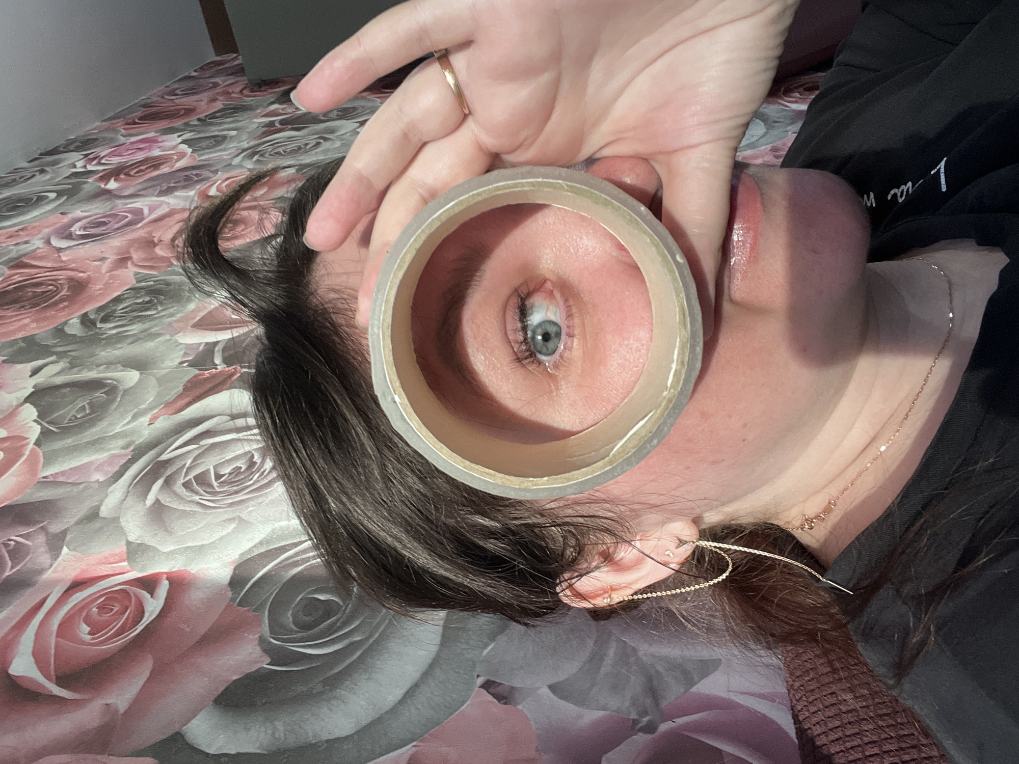 A close-up, high-angle shot captures a person lying down, looking through the cardboard core of a roll of clear adhesive tape. The person's left eye, featuring a prominent light blue-grey iris, is the central focus, framed perfectly by the tape roll. A hand with a gold ring on a finger holds the tape. The person has long, dark hair, some strands of which frame their face and ear.
They are wearing a dark garment, possibly a hoodie or sweatshirt, with white text partially visible on it, which reads "l n a." A delicate gold-colored chain necklace is visible around their neck, extending to a simple gold threader earring in their left ear. The background consists of a patterned surface, likely bedding or a blanket, adorned with large rose motifs in shades of pink, white, and grey.
The scene conveys a creative or playful moment, using an everyday object to frame the eye. There are no specific cues for time of day or weather. No other discernible text is visible besides the partial text on the garment.