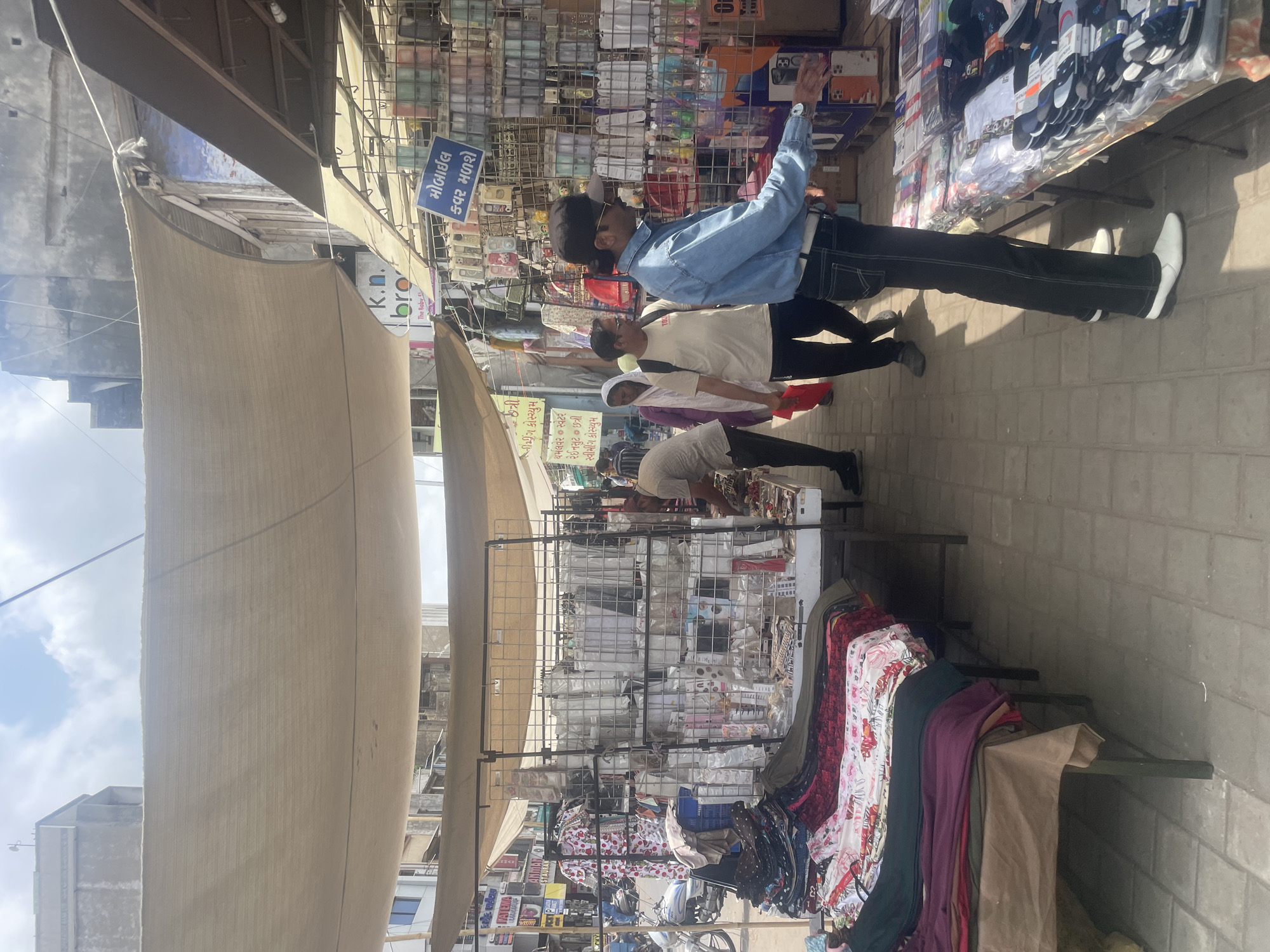 The image depicts an outdoor market scene in Ahmedabad, India, likely a busy street market. In the foreground, a stall displays folded textiles, including patterned fabrics in reds, blues, and greens. To the right, another stall is laden with various items, including packaged socks and what appear to be mobile phone cases, displayed on shelves and tables. A man in a blue denim shirt and black pants, wearing white shoes, is standing near this stall, reaching out his right hand towards the merchandise. Further into the market, a man in a light-colored t-shirt and glasses walks away from the camera, followed by a woman in a purple hijab and a red garment. Behind them, other market stalls and buildings are visible. The market is covered by a large, light-colored canvas awning. A motorcycle is parked on the left side of the image. The ground is paved with rectangular stones. The overall atmosphere suggests a vibrant commercial area with a variety of goods on display and people engaged in shopping or working.