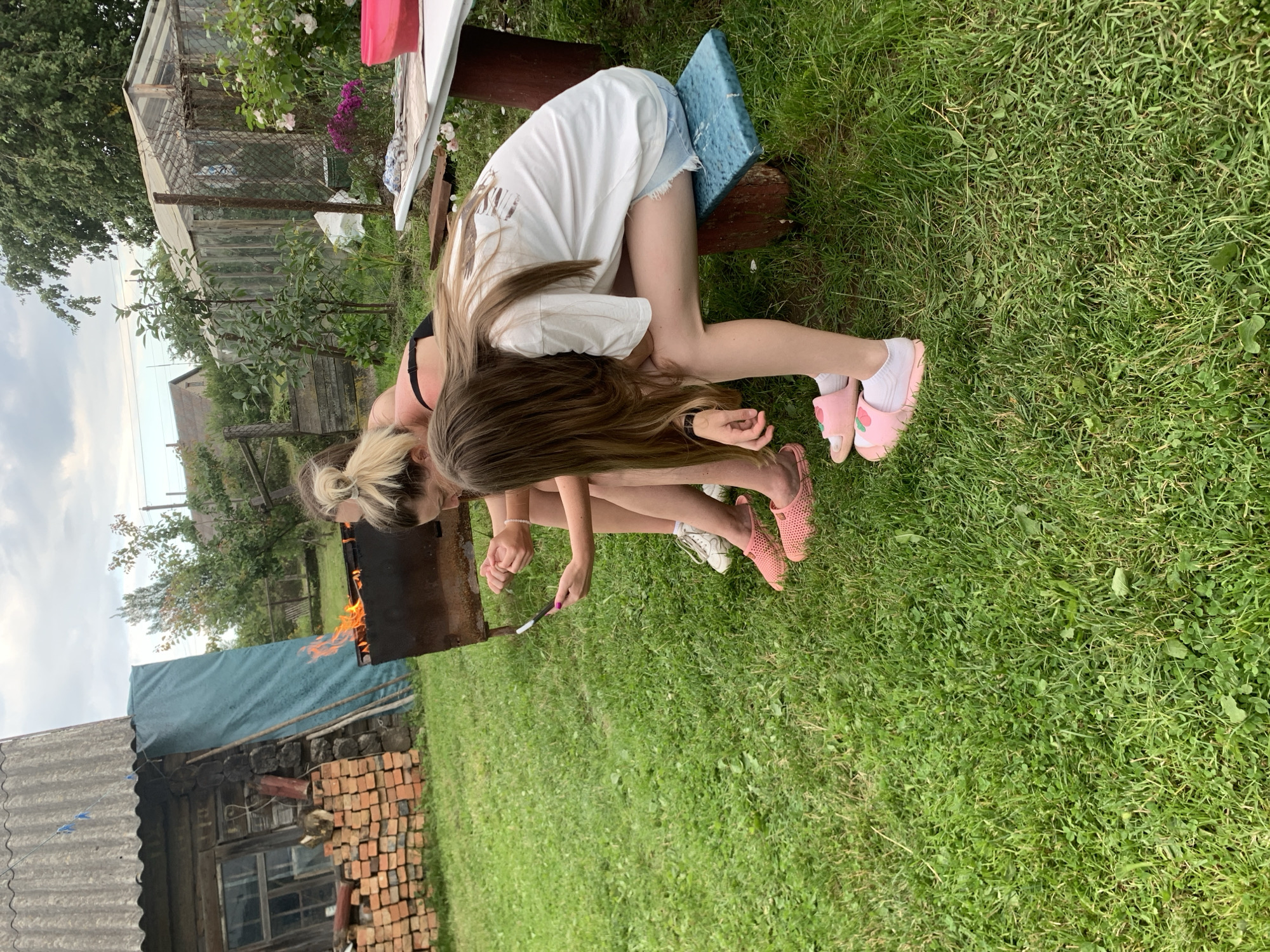 This media file depicts a scene outdoors in what appears to be a rural setting in Belarus, likely during daylight hours with overcast skies. Two young people are the primary subjects, positioned on the grass in a somewhat dynamic pose.
One individual, wearing a white t-shirt and denim shorts, is leaning forward, seated on a wooden bench with a blue object beneath them. Their hair is long and brown, falling forward over their face. The other individual, with blonde hair pulled back, is positioned lower, closer to the ground. They are wearing a dark top and appear to be interacting with a barbecue grill that has flames visible. Their legs are extended, and they are wearing pink slip-on shoes with white socks, and white sneakers.
The scene includes elements that suggest a backyard or garden area. A greenhouse structure is visible in the background, along with trees, some flowering plants, and a fence. To the left, there is a rustic wooden building with a corrugated metal roof and a stack of bricks nearby, further reinforcing the rural context. The overall mood seems casual and relaxed, possibly indicating a gathering or a moment of leisure. No visible text is present in the image.