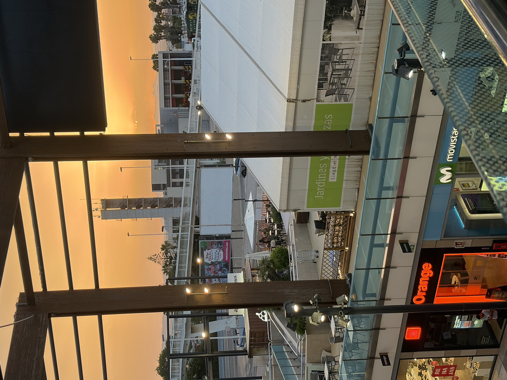 The image captures a sunset scene in Torrevieja, Spain, likely from an elevated viewpoint within a commercial or leisure complex. The sky is a vibrant orange and yellow, indicative of a sunset. In the foreground, wooden beams and metal railings of a walkway are visible. Below, a section of shops is illuminated, including an "Orange" store with its distinctive red logo and a "Movistar" store. To the right, a large white tent structure is prominent, with a green banner displaying "Jardines yazas". Beyond the tent and shops, a street with a few cars and a pedestrian overpass can be seen. Further in the background, buildings, palm trees, and a white tower structure are present. The scene appears to be in an outdoor public space, possibly a shopping center or a promenade, given the presence of retail establishments and dining areas with tables and chairs.