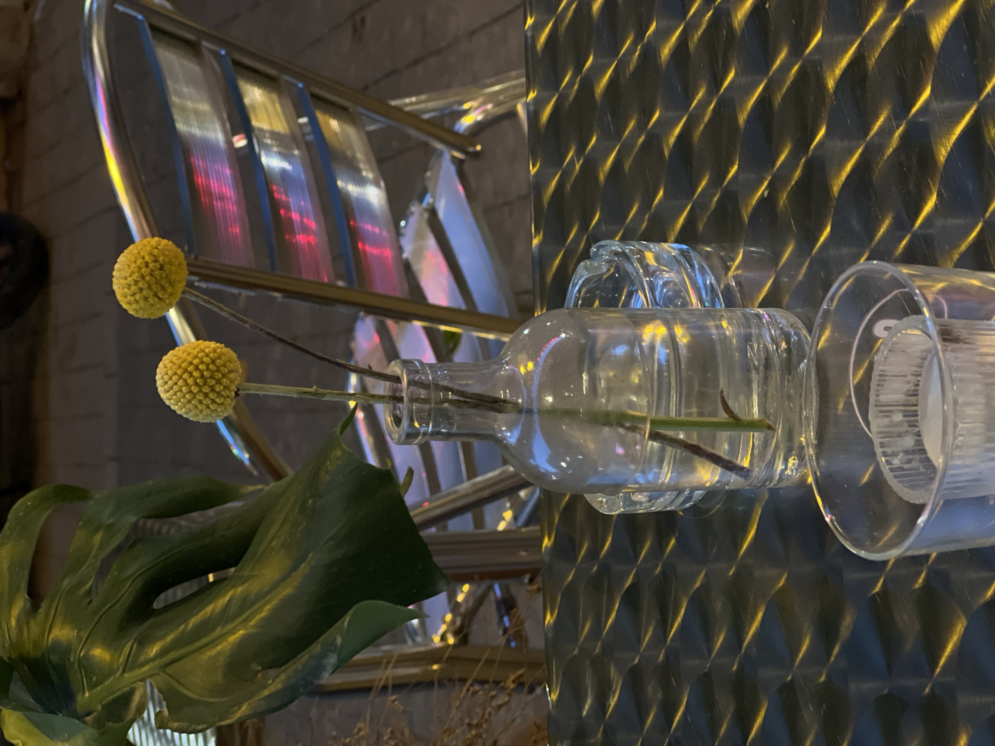 A still life scene is captured indoors, displaying a clear glass bottle lying on its side, functioning as a vase, holding two yellow Billy Button flowers and a green Monstera leaf. The bottle is partially positioned on top of what appears to be a patterned, possibly metal, wall. Beside it sits a clear, empty glass. In the background, part of a ladder with reflective rungs is visible.
While there's no confirmation, the aesthetic aligns with typical interior decoration in urban areas like Tel Aviv, Israel. However, specific details about the location are absent from the image. There are no people visible, and the focus is solely on the arrangement of the objects.