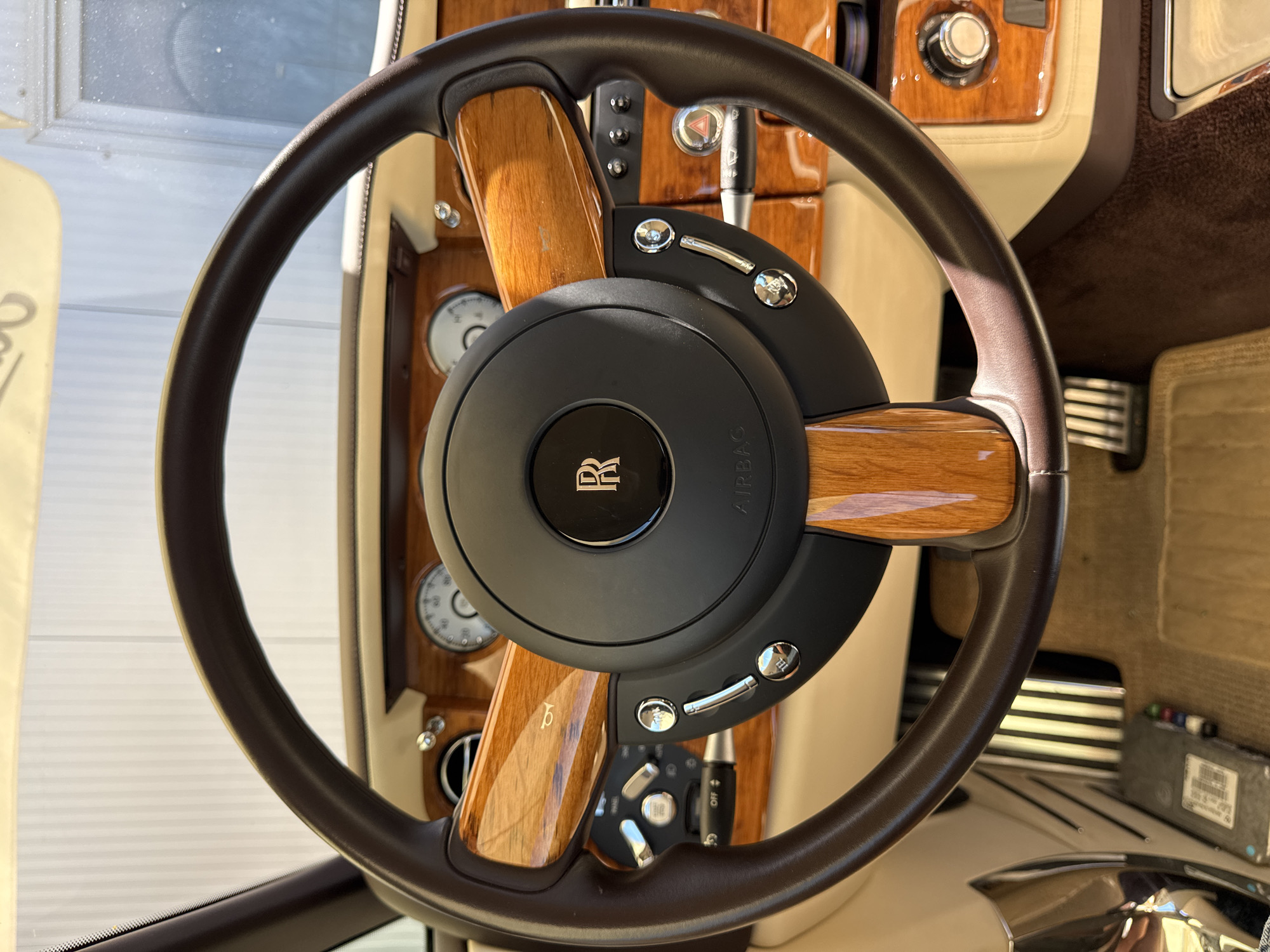 The image shows a close-up of a luxurious car's steering wheel and dashboard. The steering wheel is made of dark brown leather with polished wood accents. In the center of the steering wheel is the Rolls-Royce logo, a stylized "R". The dashboard is adorned with more polished wood trim and chrome accents. There are also various buttons and controls visible. The interior of the car appears to be very well-maintained and opulent. The location in the city of Dub, Montenegro, is not discernible from the image.