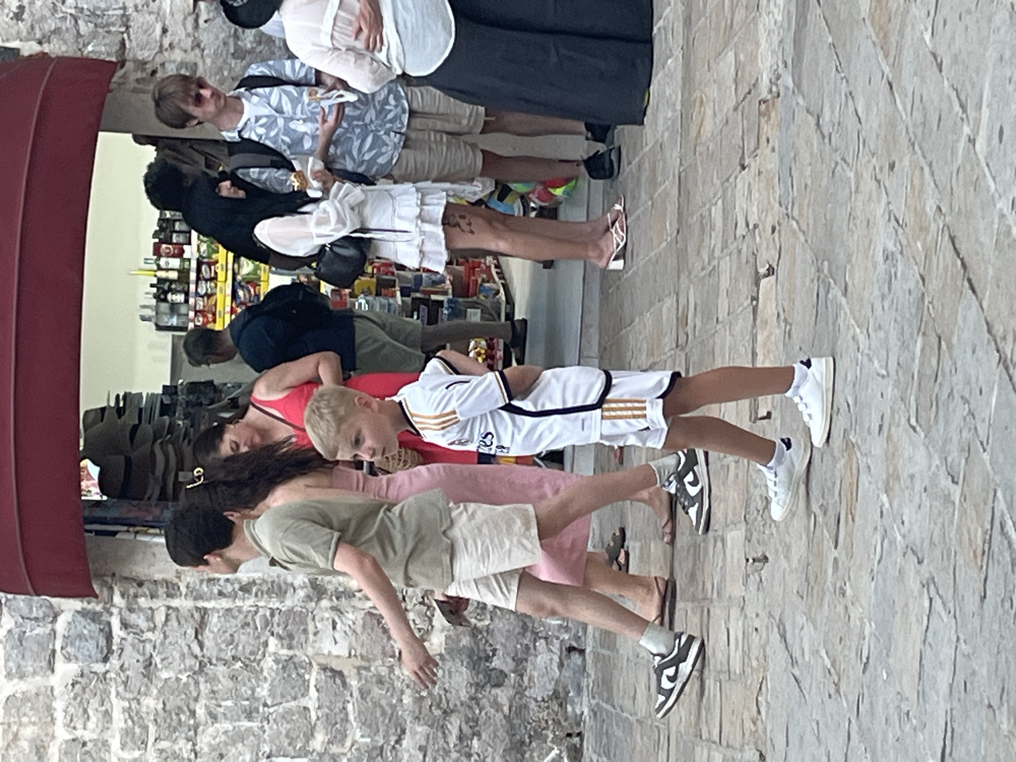 In the city of Budva, Montenegro, a group of people are walking on a cobbled street in front of a building with a stone facade and a red awning. A young boy in a white soccer uniform walks in the foreground, looking to the right. Behind him, a man in a green polo shirt and beige shorts walks from left to right, his left arm extended. To his right, a woman in a pink dress and another woman in a white dress are partially visible. Further back, a man in a blue patterned shirt and beige shorts stands and holds a sandwich, while a woman in a black dress and a woman in a white top and dark skirt are also present. A store with shelves of merchandise is visible in the background.