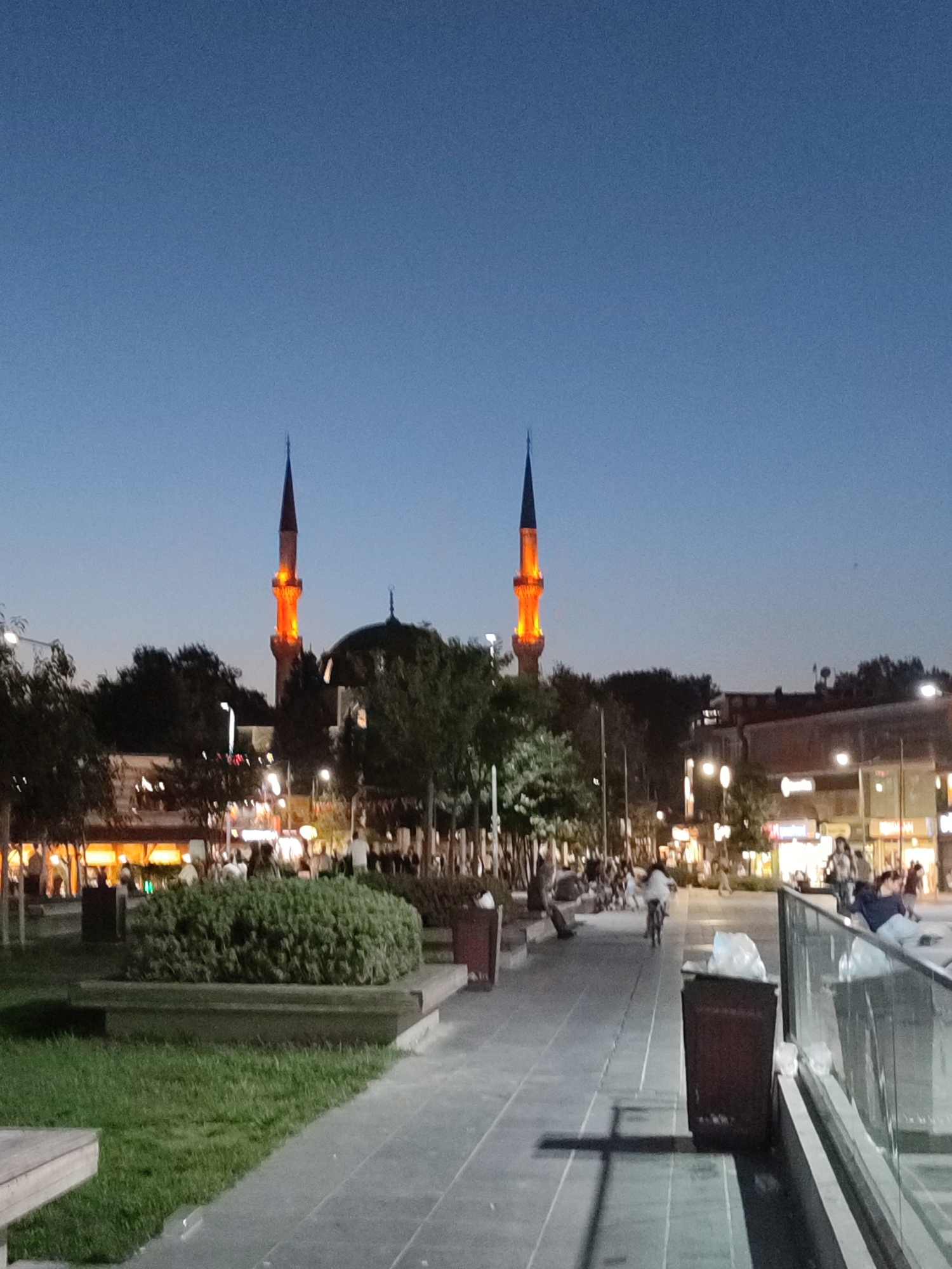 This is a vertical, full-shot, low-angle, outdoor, eye-level, wide-angle photograph of a city street scene at dusk in Üsküdar, Turkey. Two minarets of a mosque are illuminated with orange light, standing prominently against the clear, darkening blue sky. A dome of the mosque is visible between the minarets. The street is paved with gray stones and has a sidewalk lined with a low, green hedge and grass. Several trees are scattered throughout the scene. In the background, buildings with illuminated windows and storefronts line the street, indicating shops and businesses. A glass railing runs along the right side of the sidewalk, with trash cans visible nearby. Several people are present in the scene, walking, sitting, and one person is riding a bicycle. The overall atmosphere is that of an active urban environment at the end of the day.