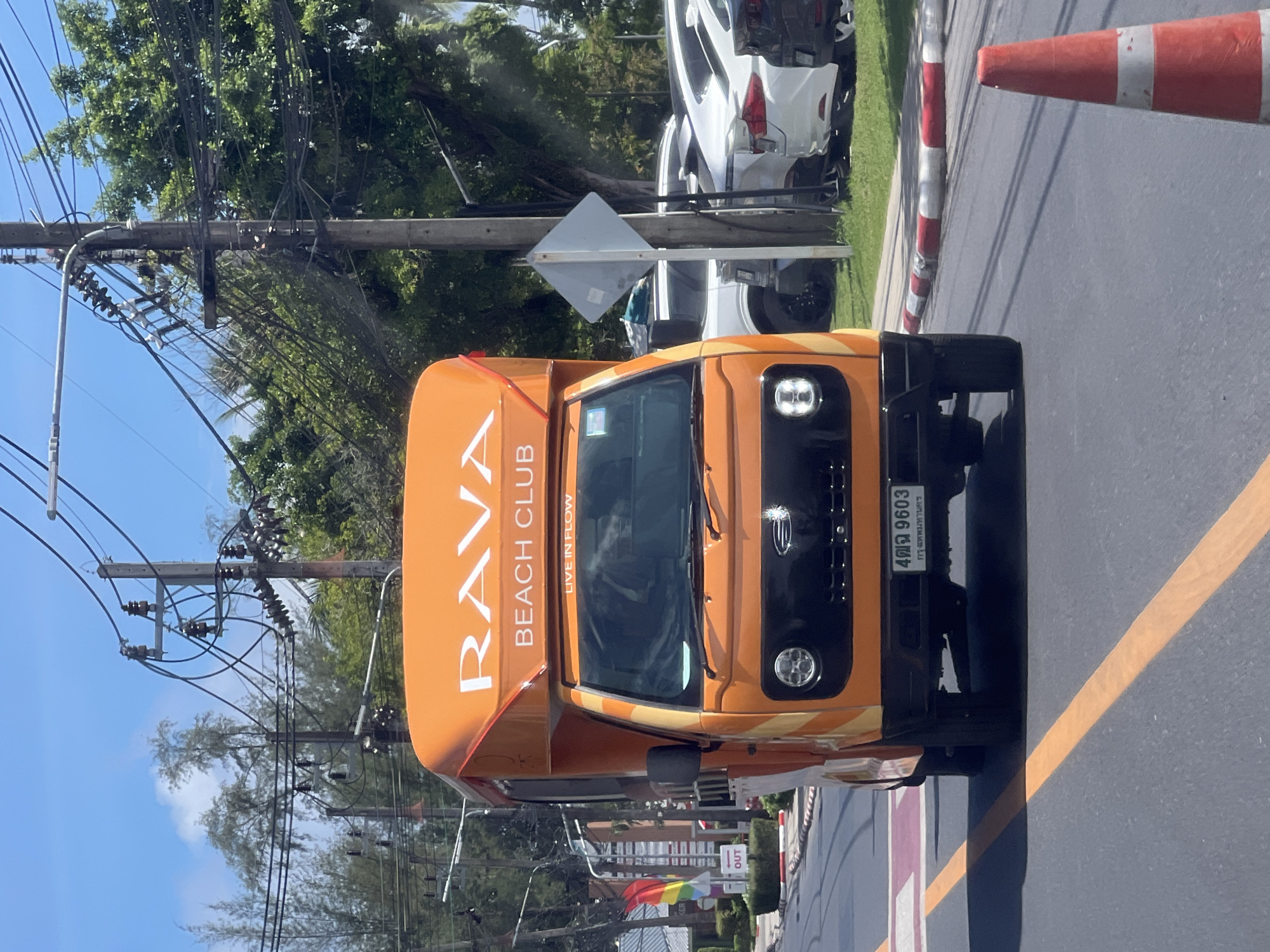 An orange truck with "RAVA BEACH CLUB LIVE IN FLOW" written on the side is driving on a street. The truck has a license plate that reads "4 9603". In the background, there are trees, power lines, and parked cars. A traffic cone is visible on the side of the road.