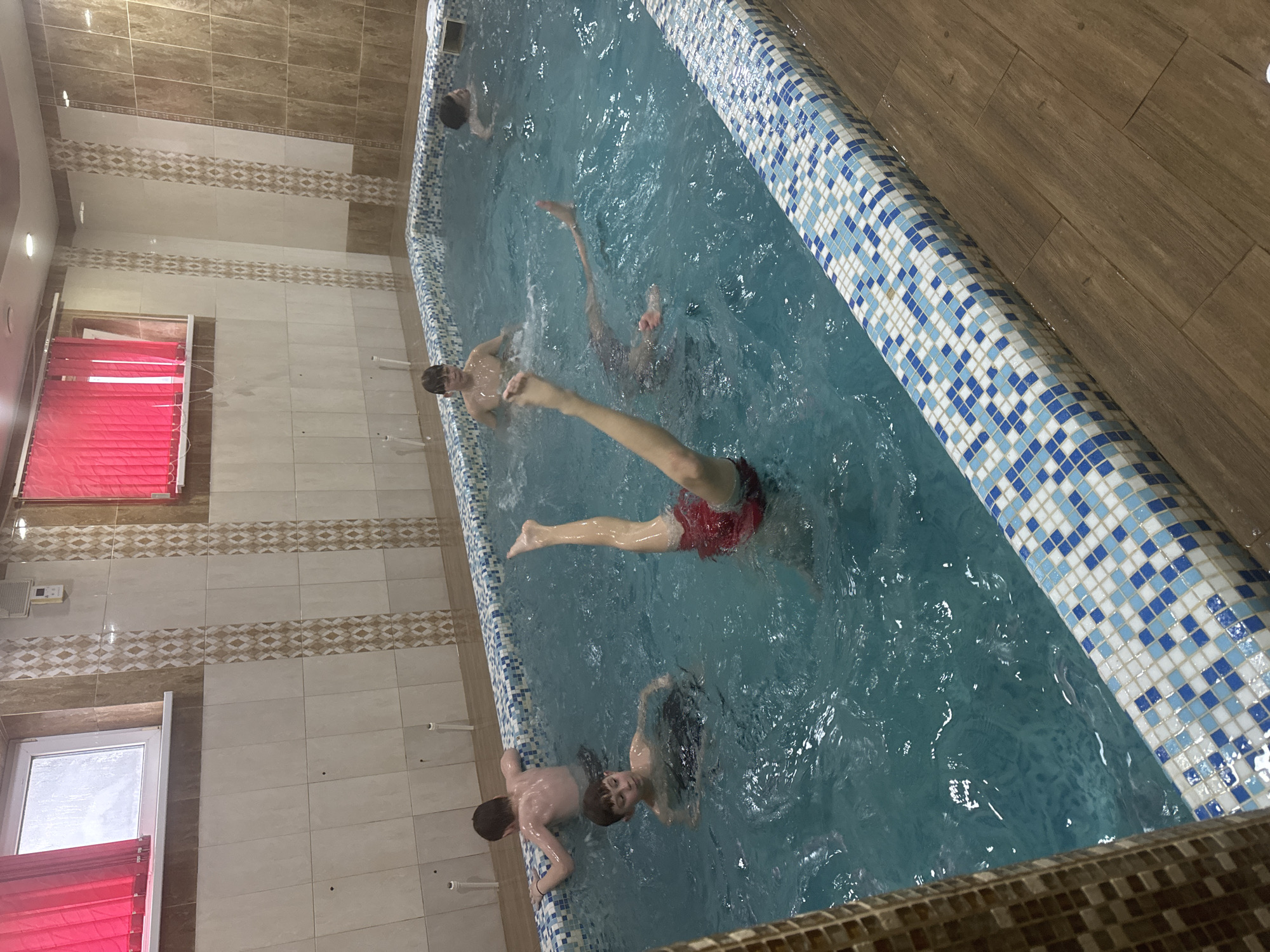 This image depicts an indoor swimming pool scene in Chaikovsky, Russia, where several young males are present. The pool features blue and white mosaic tiles along its edge, filled with light blue, agitated water. The surrounding floor is covered with brown, wood-patterned tiles, while the walls are adorned with light beige rectangular tiles and decorative horizontal bands of brown and beige patterned tiles.
At least five individuals, appearing to be boys or young men, are in the pool. In the foreground, one person wearing red swimming shorts has their legs extended above the water. Near the left side of the pool, another individual leans against the edge. Towards the center, one person's head and shoulders are visible above the water, looking towards the camera, while another's upper body is partially submerged nearby. A fifth individual is partially visible at the far end of the pool.
On the left wall, a window is covered with bright red Venetian blinds. Above the blinds, a small white rectangular object, possibly a control panel or thermostat, is mounted. Ceiling lights illuminate the indoor environment. The scene portrays a recreational activity, with the individuals actively engaged in the water. No visible text is present in the image.
