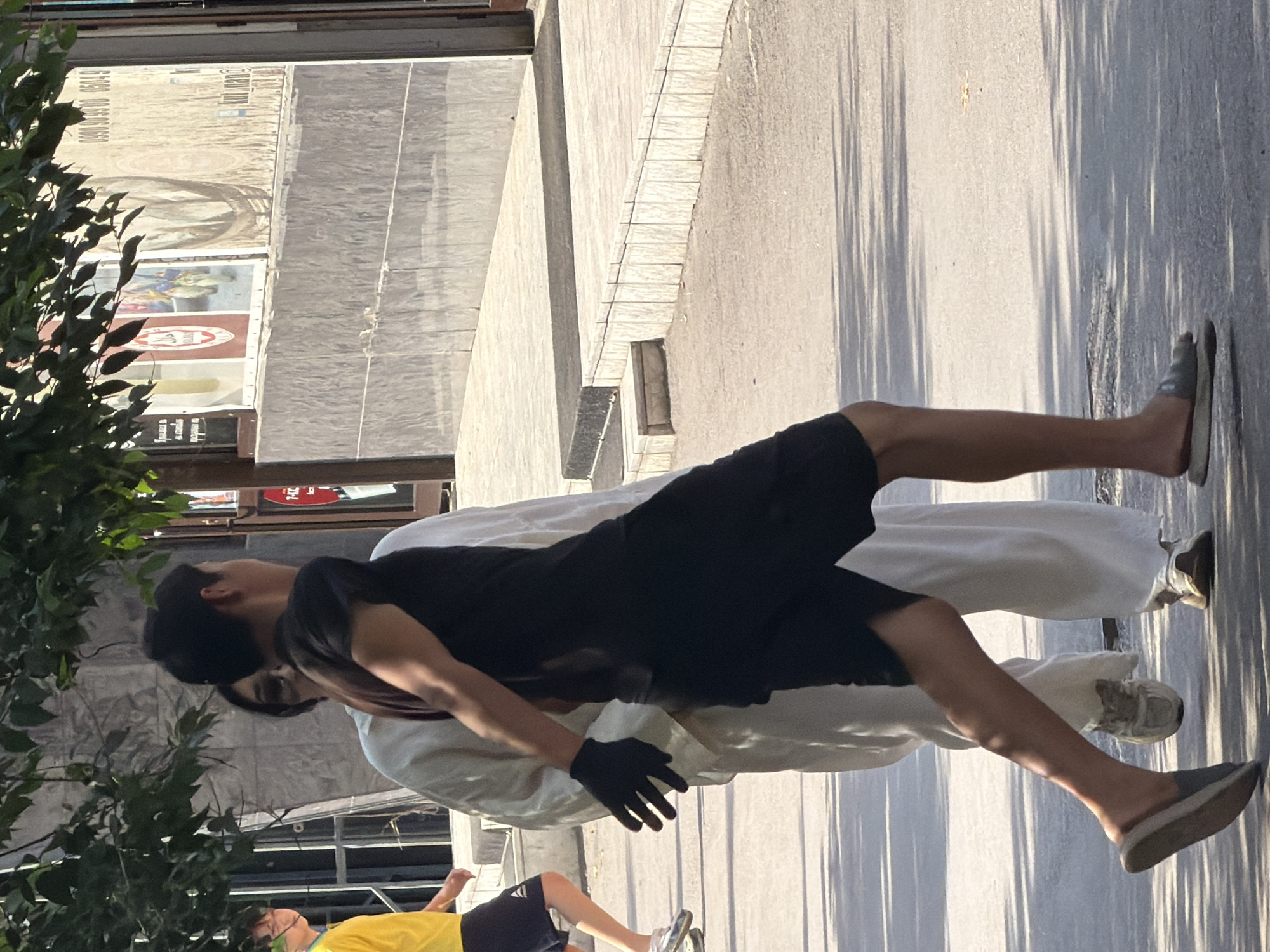 In the city of Unknown, Unknown, two individuals are walking on a sidewalk. The person in the foreground, wearing a black t-shirt and black shorts, is moving from left to right, with their right arm extended and gloved. They are wearing a dark-colored sandal on their left foot and a lighter-colored sandal on their right foot. To their left, another individual is wearing a long white garment, partially obscuring their face, and appears to be walking in the same direction. Further to the left, a person in a yellow t-shirt and dark shorts is walking away from the camera. The environment suggests an urban setting with a tiled wall and building facades in the background, partially covered by leafy branches overhead. The ground is paved with concrete and asphalt.