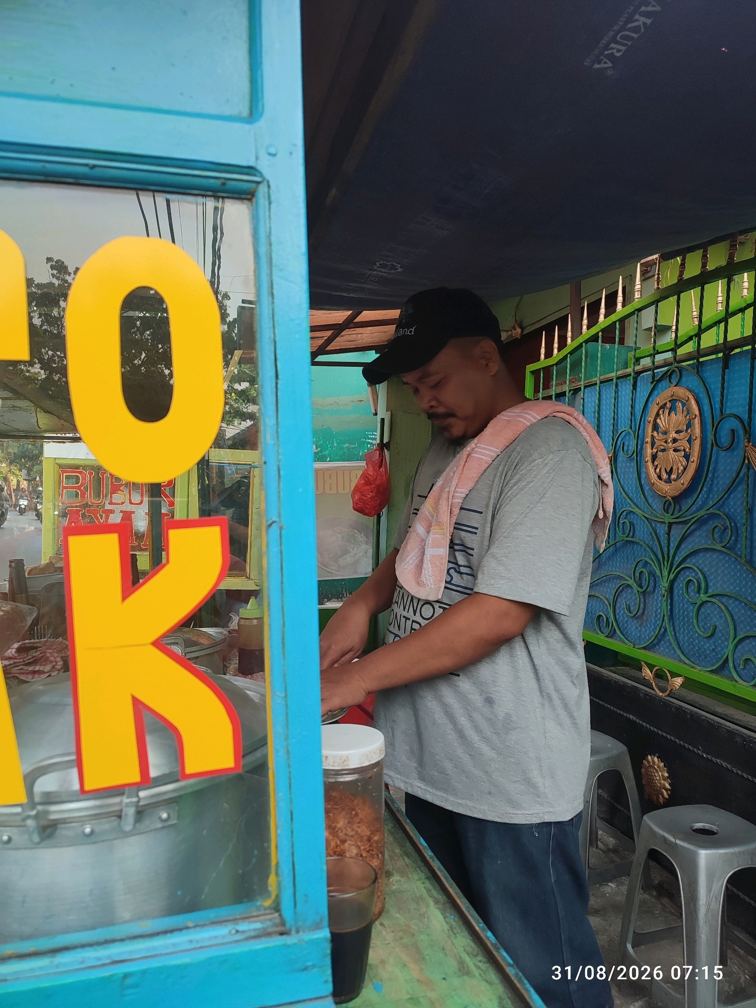 A medium shot captures a man standing behind a food cart in East Jakarta, Indonesia, at 7:15 AM on August 31, 2026. The man, wearing a gray t-shirt, dark blue jeans, and a black baseball cap, has a towel draped over his shoulders. He is engaged in food preparation, with his hands in the process of working. The food cart is adorned with large yellow letters, and its glass window offers a view of the street with passing motorcycles and trees. In the background, a blue metal fence with decorative elements and green accents is visible, along with two gray stools. A tarp is stretched overhead, providing shade.