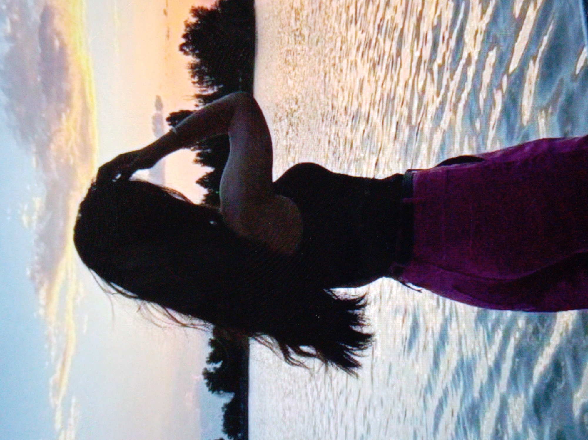 The image depicts a person in silhouette with long, dark hair, leaning against a body of water. The person is wearing dark clothing on their upper body and a purple garment on their lower body, possibly shorts or a skirt. The background consists of a body of water with ripples and reflections, and a sunset or sunrise in the sky, casting warm hues of orange and yellow. Trees are visible on the left side of the image, silhouetted against the sky. The perspective is from a low angle, looking up towards the person and the sky. The image appears to be taken outdoors, likely near a lake or ocean during twilight. The location is Unknown, Unknown.