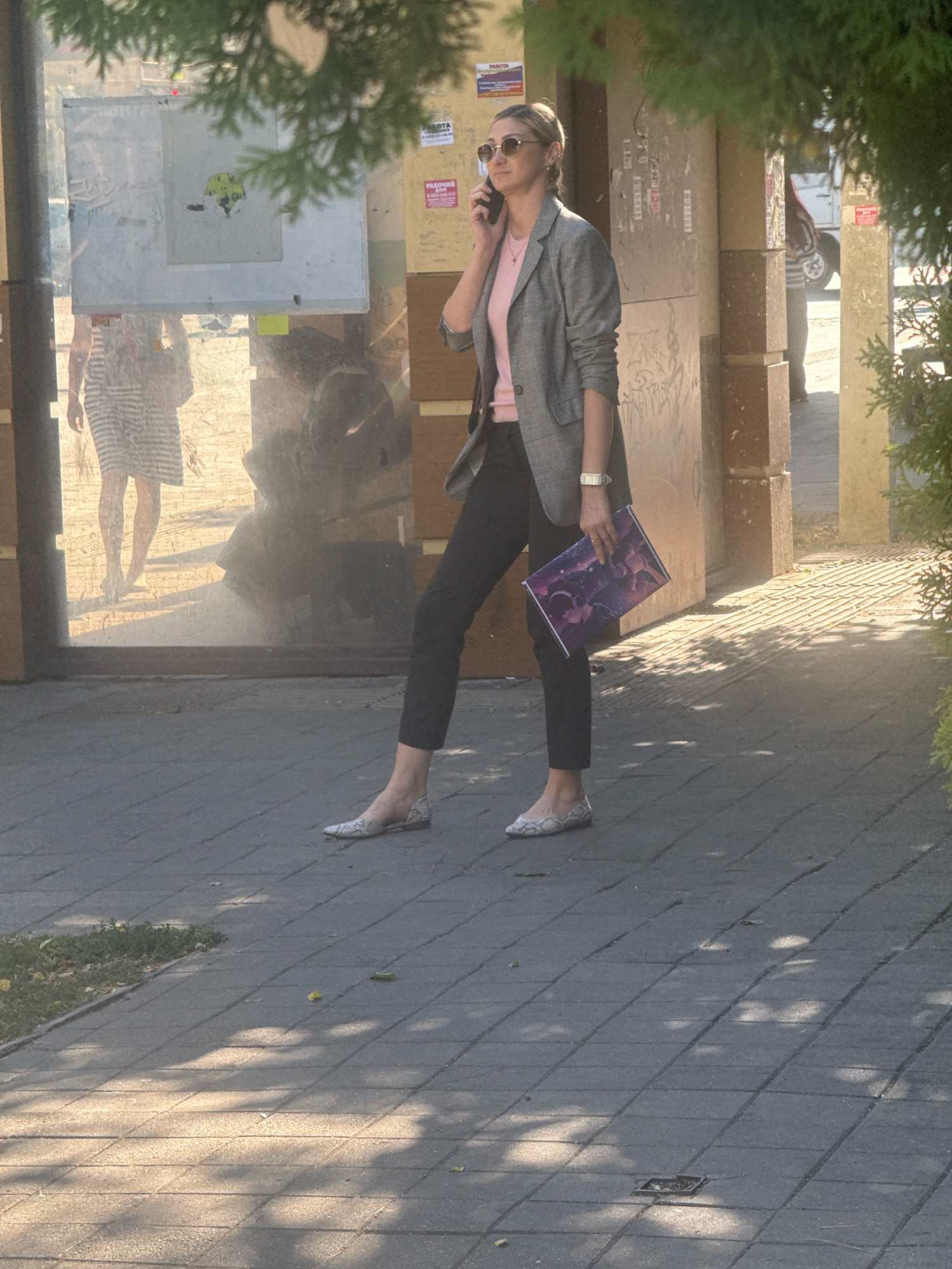 A woman wearing a grey plaid blazer, a pink t-shirt, and black pants stands on a sidewalk. She has blonde hair pulled back and wears sunglasses. She holds a cell phone to her left ear with her left hand and a folder with an image of a nebula or galaxy with her right hand. She wears flat, white, patterned shoes. A reflection in a large glass pane behind her shows another woman in a striped dress. The sidewalk is paved with grey interlocking bricks and has shadows cast from trees. The environment appears to be an urban setting, possibly in Anapa, Russia, based on context clues from the provided information about the city.