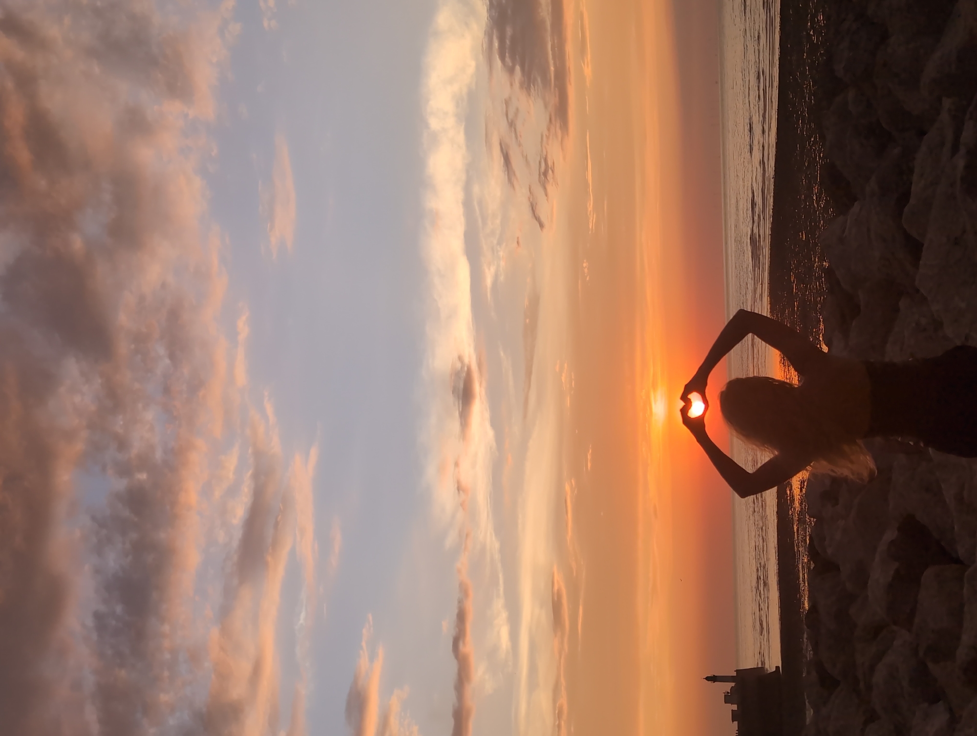 A silhouette of a person stands on a rocky shore, facing the sea during sunset. Their arms are raised, and their hands form a heart shape around the setting sun, which is partially obscured by clouds. The sky is painted with hues of orange, pink, and purple, with scattered clouds reflecting the warm light. In the distance, a lighthouse stands on a pier, and the calm sea stretches to the horizon. The foreground is dominated by large, grey rocks. This scene appears to be captured at Le Tréport, France, a coastal town known for its picturesque sunsets over the English Channel.