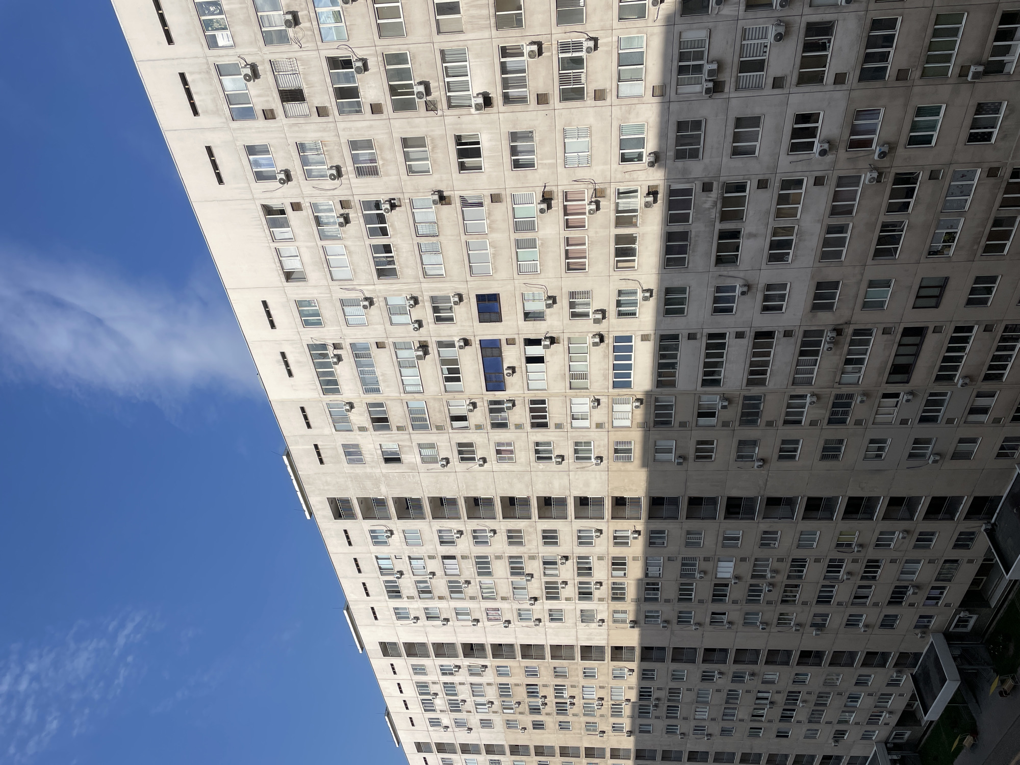 The image shows a long, multi-story apartment building in Almaty, Kazakhstan. The building is constructed from concrete panels and is characterized by numerous windows and air conditioning units protruding from the facades. A shadow from an unseen object falls across the right side of the building, creating a stark contrast between the sunlit and shadowed sections. The sky above is blue with some scattered white clouds. The lower left portion of the image reveals a small section of the surrounding urban environment, including a covered walkway, grass, and what appear to be benches. No people are visible in the image. The architectural style suggests a Soviet-era mass housing project, common in many post-Soviet cities like Almaty.