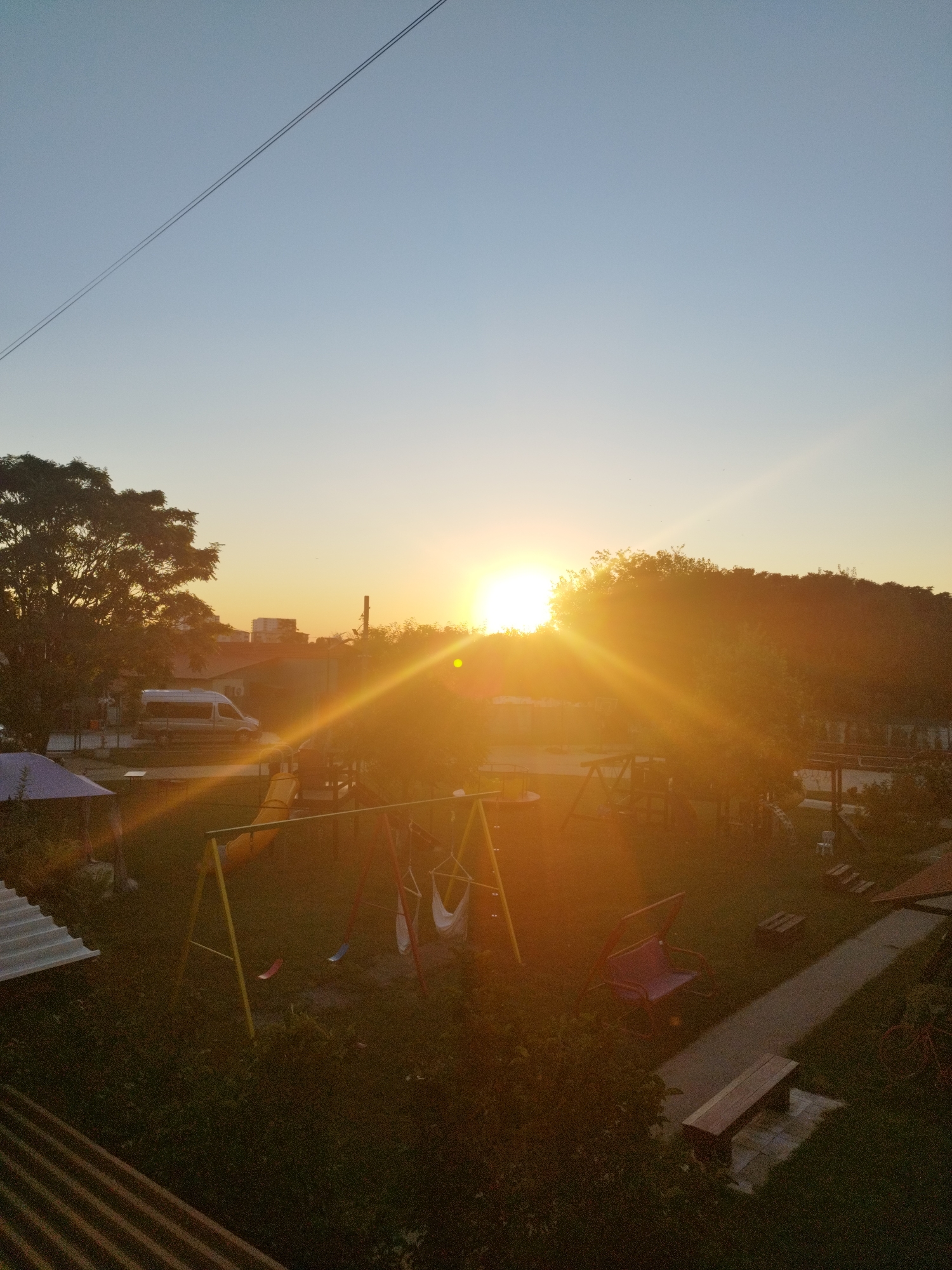 The image depicts an outdoor scene at sunset, with the sun prominently positioned on the right side of the frame, casting a bright orange glow and lens flare across the entire image. In the foreground, there are playground elements including a yellow slide, swings with red and blue seats, and a hammock-style swing. Adjacent to the playground, there is a blue-roofed structure and what appears to be a white corrugated metal roof. Further back, a paved walkway is visible, with a wooden bench situated next to it. Other elements in the background include trees, buildings, and a white van. The sky transitions from a clear pale blue at the top to a warm orange hue near the horizon. A power line stretches diagonally across the upper portion of the sky. The scene is likely a residential area or a park, possibly in Bucharest, Romania, given the architectural style of the buildings and the general ambiance. There are no people visible in the image.