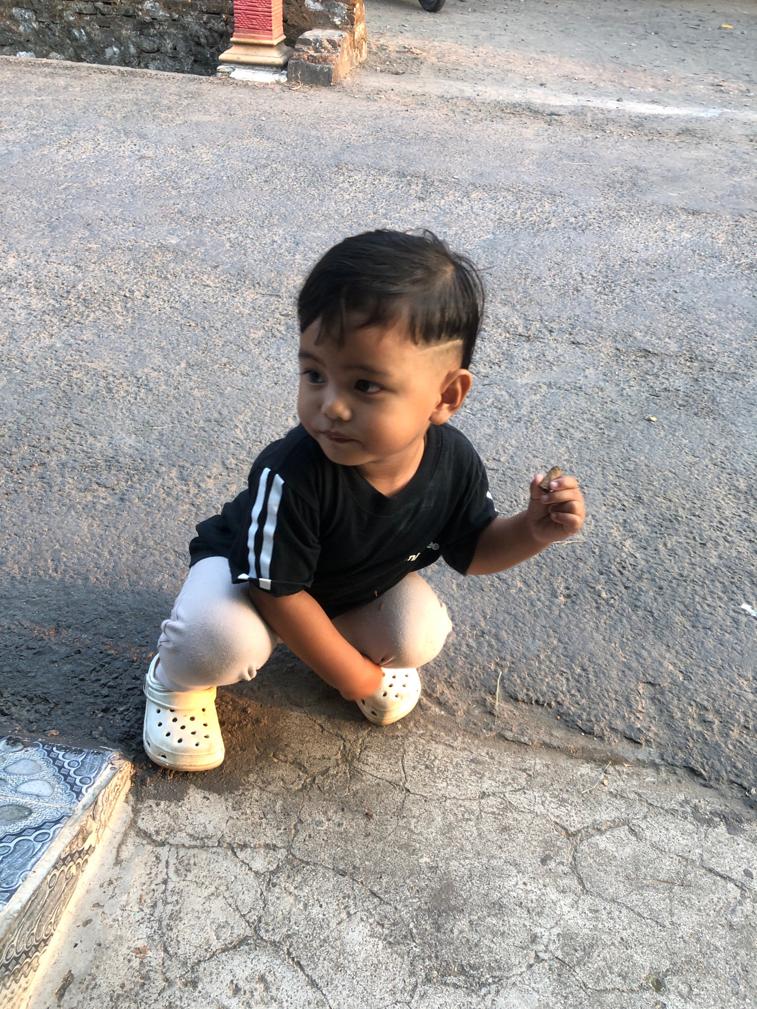 A young child, approximately two to three years old, is crouching on a paved surface in what appears to be an outdoor area of Banjarejo, Indonesia. The child has dark hair, styled with a distinct fade on the sides, and is wearing a black t-shirt with white stripes on the sleeves and light-colored pants. They are also wearing white Croc-style shoes. The child is looking to their left and holding a small object, possibly an insect or a piece of debris, in their right hand. The ground is asphalt with some cracks and patches of concrete. In the background, there is a stone wall with a decorative red and yellow pillar, and a hint of what might be a building or structure. The lighting suggests it is daytime, possibly late afternoon, with shadows cast by the child. The overall environment appears to be humble and possibly rural or semi-urban.
