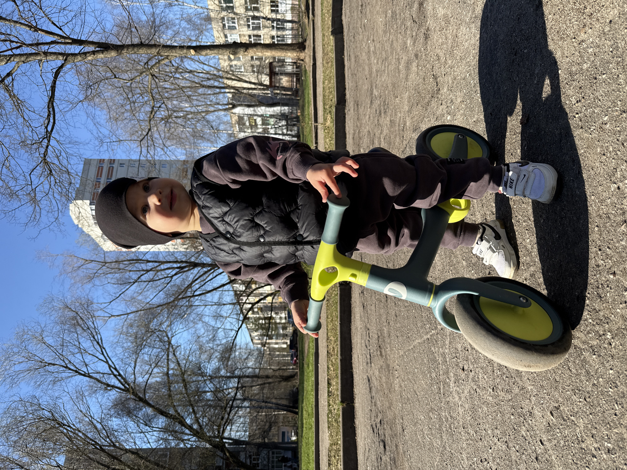 A young child wearing a black cap, dark brown jacket, and white Nike sneakers is riding a light blue and yellow tricycle on a paved surface. The child is looking towards the camera. The background includes bare trees, grass, and apartment buildings. The scene is Rusanovo, Russia.