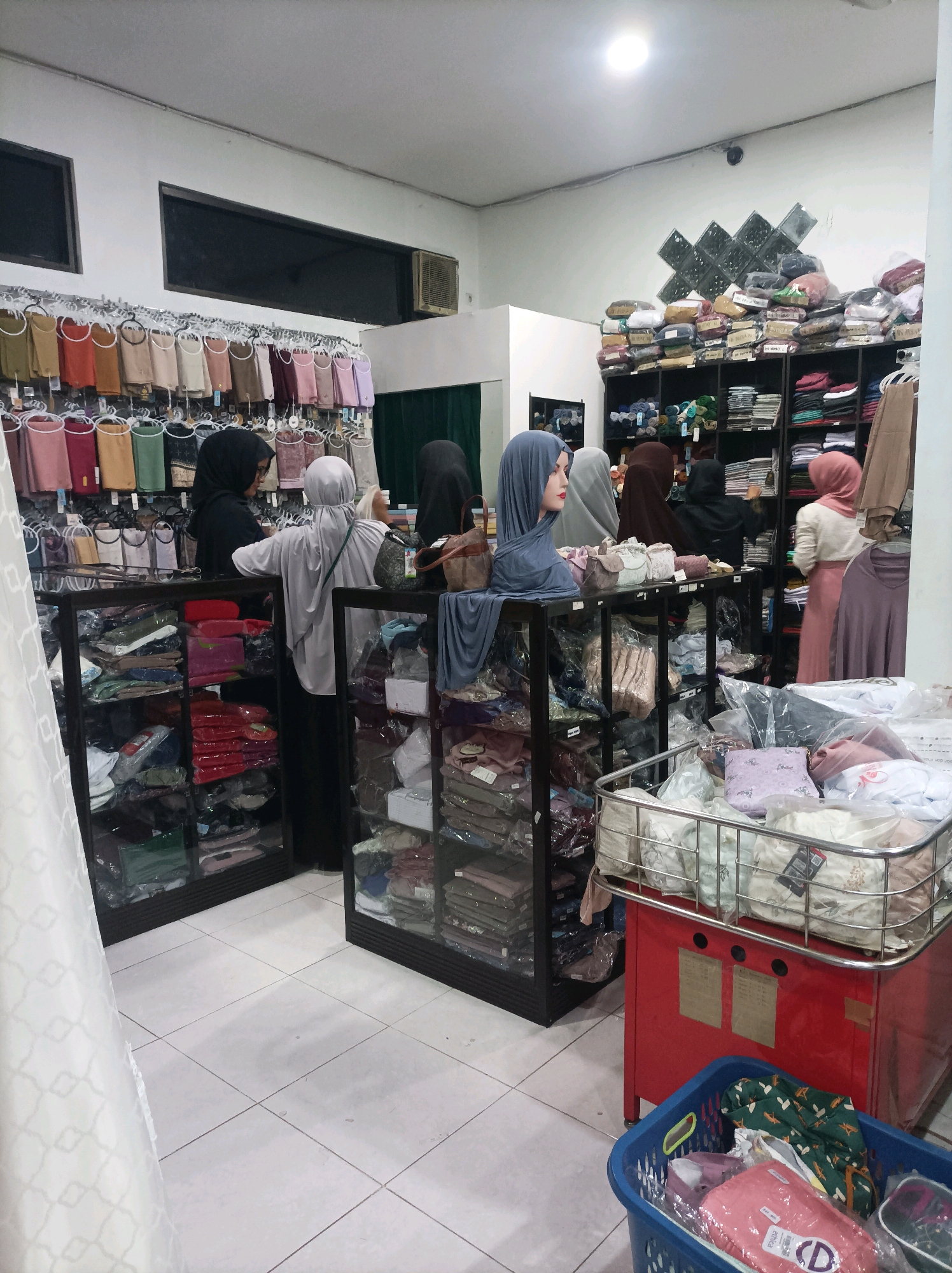 The image shows the interior of a clothing store, likely in Blimbing, Indonesia, specializing in Islamic attire. The store is filled with shelves and display racks containing various types of hijabs, abayas, and other modest clothing items. Several women, some wearing hijabs, are browsing the merchandise. Mannequins displaying hijabs are also visible. The store appears to be well-stocked, with clothing organized by color and type. The lighting is bright, indicating an indoor commercial setting. A checkout counter with a red cabinet and a blue basket filled with clothes is also present in the foreground.
