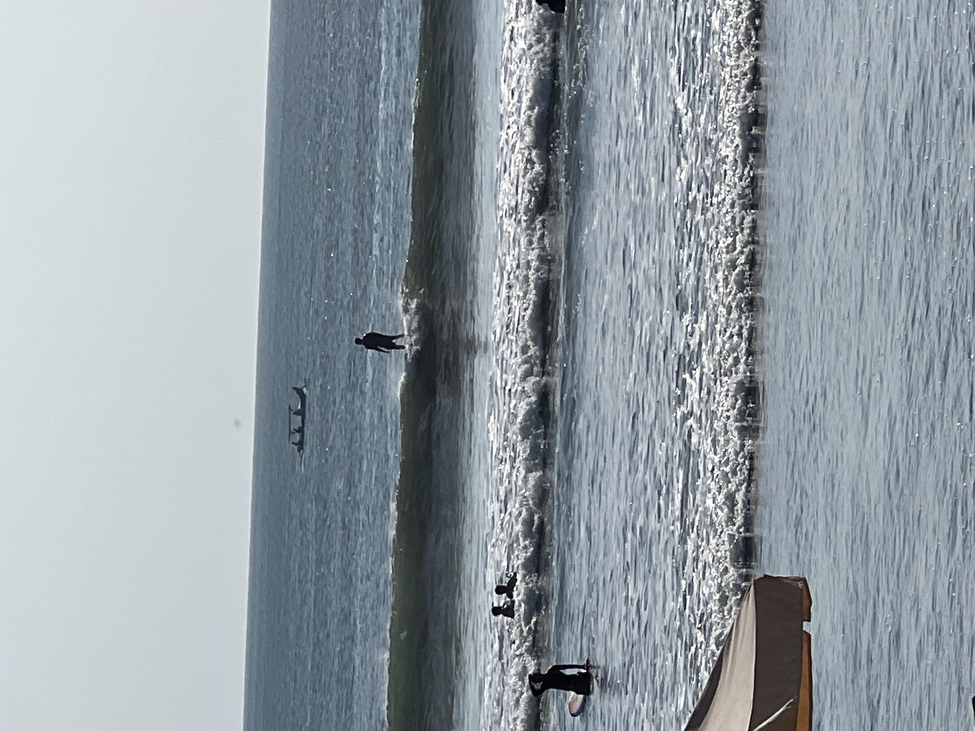 The image shows a hazy view of the ocean near Kuta, Indonesia, on a bright, overcast day. Several surfers are scattered across the water. In the foreground, a surfer sits on a surfboard facing away from the camera, with their hand raised to their head. Further out, two figures are in the water, and a single surfer is riding a wave. In the distance, a small boat with several passengers navigates the choppy sea. A portion of a tan beach umbrella is visible in the lower left corner.