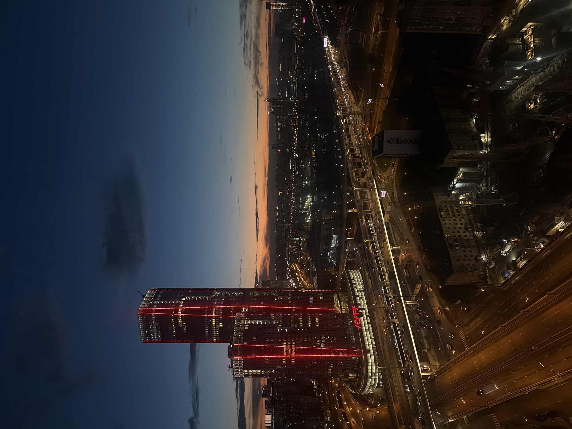 This is a nighttime aerial photograph of the city of Khimki, Russia, specifically focusing on the "I CITY" complex. The sky is a deep twilight blue, transitioning to orange and yellow hues near the horizon, indicating either sunrise or sunset. Prominently featured is a pair of illuminated skyscrapers, with red LED lighting accentuating their vertical edges and architectural features. The lower portion of the complex features a curved building with visible signage that reads "I CITY". Surrounding the skyscrapers is a complex network of multi-level highways and overpasses, filled with the headlights and taillights of numerous vehicles, suggesting active traffic. In the foreground, a multi-lane highway stretches out, illuminated by streetlights and the glow of passing cars. Beyond the illuminated highway, residential buildings and construction sites are visible, some with lights on inside. Construction cranes are scattered throughout the scene, indicating ongoing development. The overall impression is an urban landscape at dusk, characterized by modern architecture, busy infrastructure, and the beginnings of nighttime illumination.
