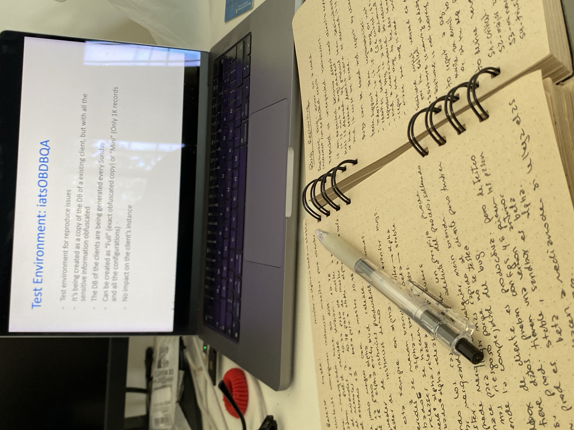 A laptop displaying a presentation titled "Test Environment: iatsOBDBQA" sits open on a desk. The presentation outlines the purpose and characteristics of the test environment, including its creation from a client's database, sensitive information obfuscation, weekly generation, and the option for full or mini records. Below the laptop, a spiral-bound notebook with handwritten notes is open, featuring sections on "Bug Reporting" and other details. A pen rests on the notebook. The background shows a blurry view of a desk with some cables and other indistinct objects. The scene appears to be an indoor workspace, with no visible connection to Villa Carlos Paz, Argentina.