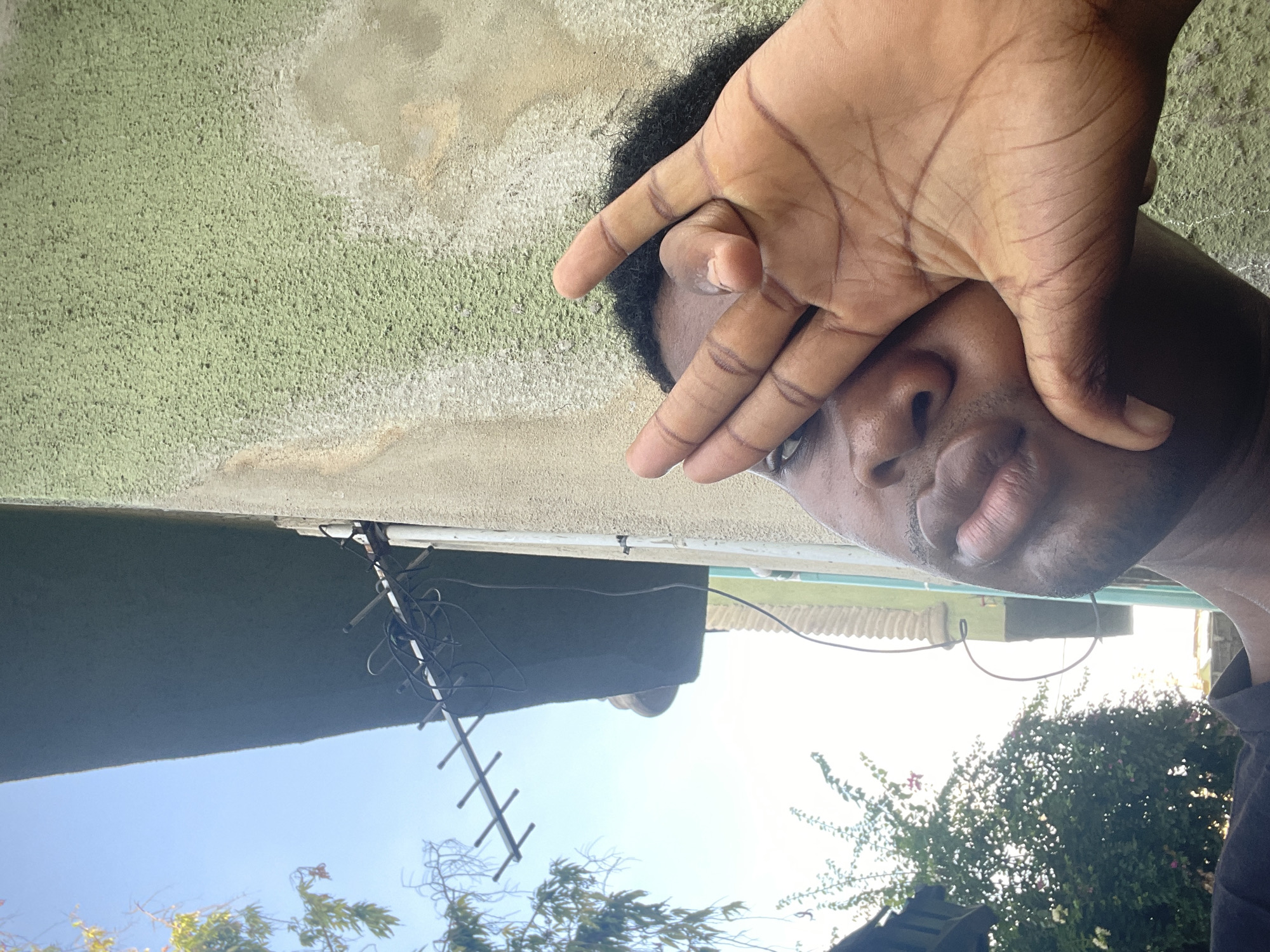 A dark-skinned person with dark hair is shown partially covering their face with their hand. The person appears to be outdoors. Behind the person, there is a light green textured wall, the corner of a building, a television antenna, part of a white corrugated metal structure, trees, and a blue sky. Due to the lack of identifying details, it is not possible to verify that the location is Ado, Nigeria.