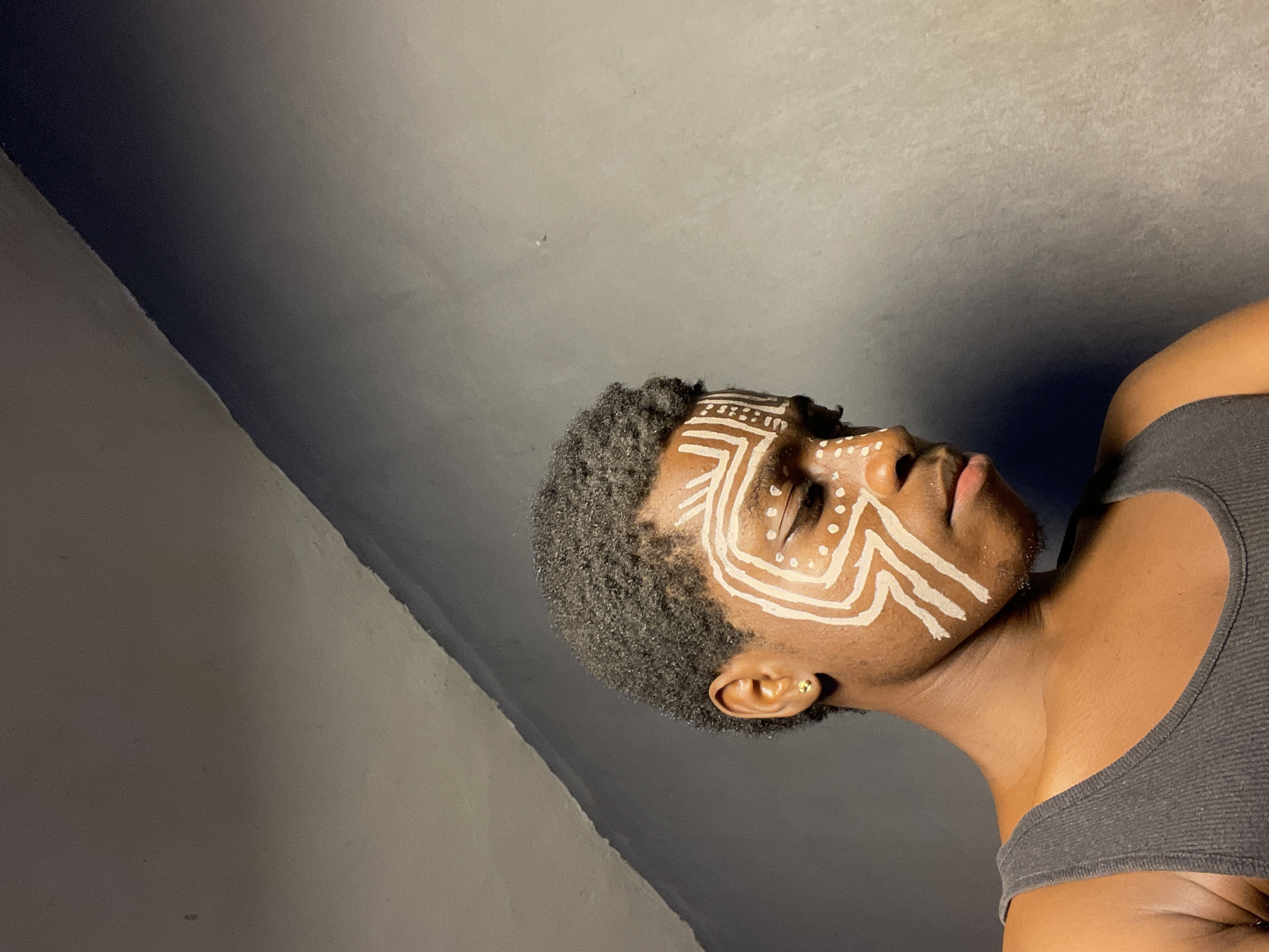 A medium-shot, canted angle photograph captures a dark-skinned person with short, dark hair, adorned with white painted designs on their face resembling tribal markings. The person is wearing a gray tank top. The background consists of two wall sections in varying shades of gray. The image was most likely taken indoors in the city of Nkpor, Nigeria. There are no discernible objects or interactions present.