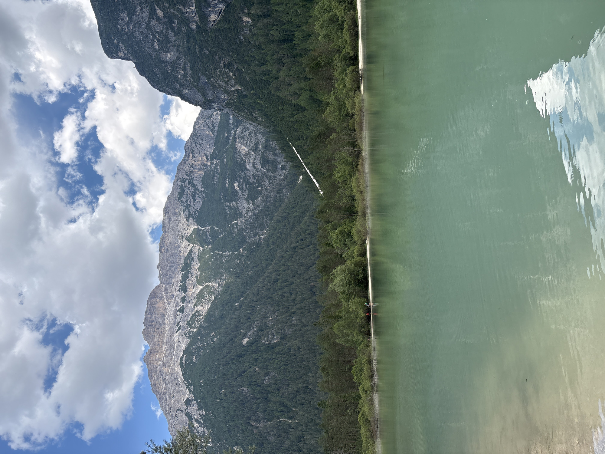 This is a landscape photograph of a clear, turquoise lake with mountains in the background. The lake occupies the lower half of the image and reflects the sky and surrounding trees. On the shore of the lake, two small figures are visible, appearing to stand near each other. Beyond the lake, a dense forest of green trees covers the slopes of the mountains. The mountains themselves are rocky and imposing, with patches of snow or light-colored rock visible near their peaks. The sky above is partly cloudy, with bright white clouds against a blue background. The overall scene depicts a serene and natural environment. The location is not specified within the city of Unknown, Unknown, but it is likely a natural park or a scenic area.