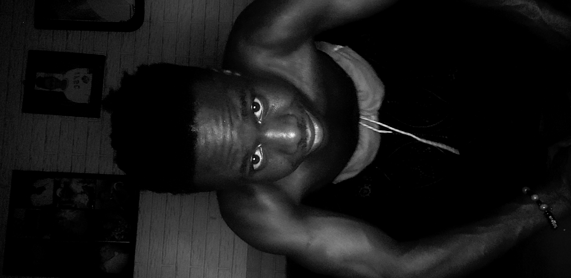 A black and white photograph depicts a dark-skinned man with short, dark hair looking upward at the camera. He is wearing a tank top with drawstrings. The man's right arm is visible, and a beaded bracelet is on his wrist. In the background, on the left side of the frame, hang two framed pictures on a wall. This photograph was taken indoors, in Narayi, Nigeria.