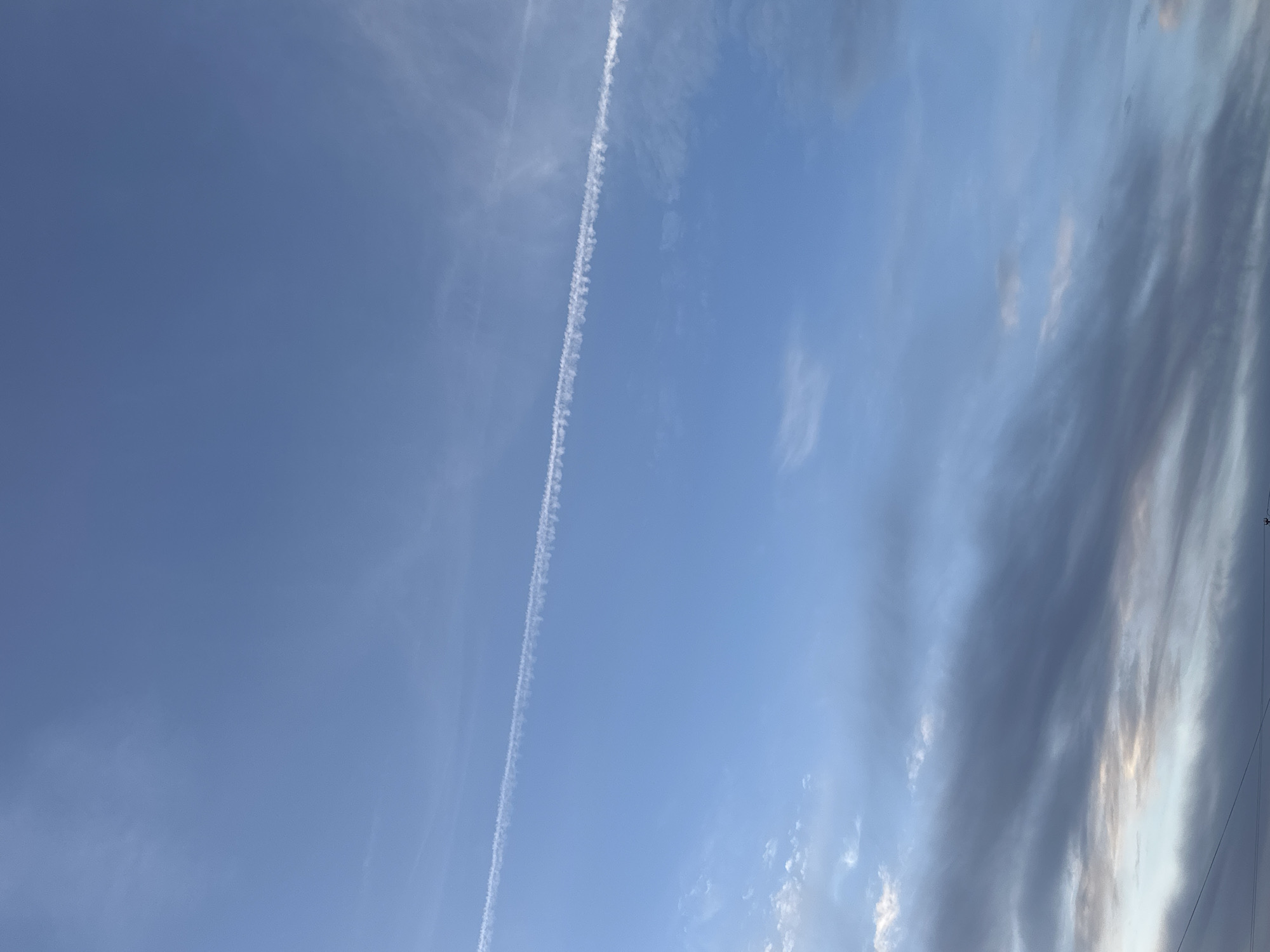 The image shows a daytime sky in Chaikovsky, Russia. The sky is mostly blue, with some areas of clouds on the right side and contrails from aircraft cutting across the blue expanse.
