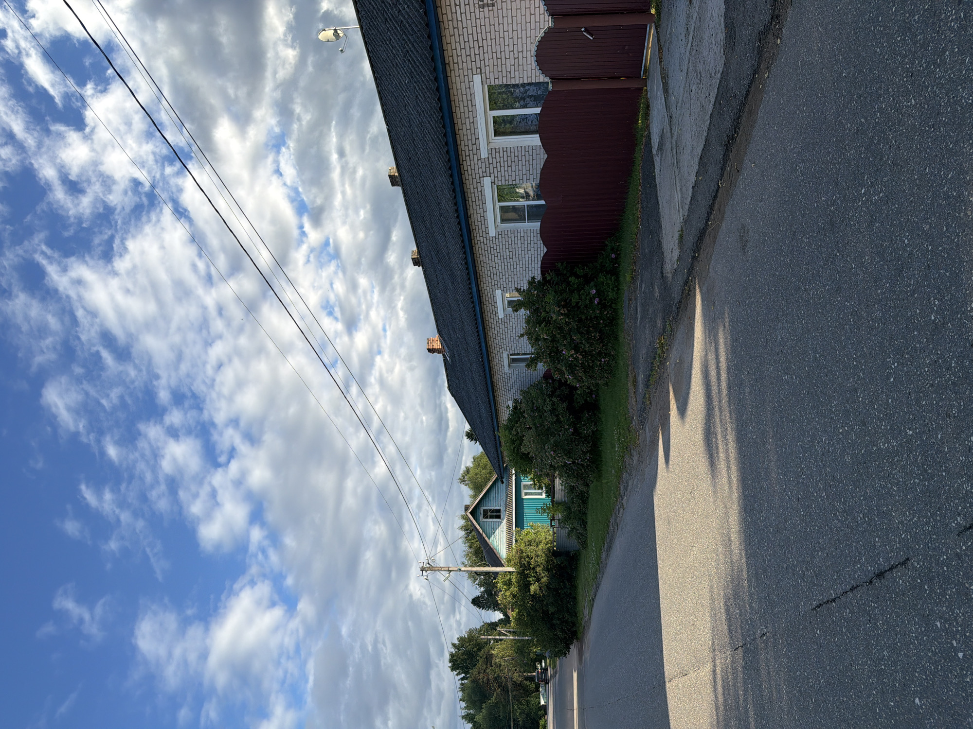 The image depicts a roadside view in Moshenskoe, Russia, under a partly cloudy sky. The scene is oriented vertically, with the road in the foreground and buildings and sky in the background.
The main subjects are two houses, a paved road, and a sky filled with dramatic clouds. The house closest to the viewer is a light-colored brick building with a dark roof and a reddish-brown wooden fence. It has several windows with white frames. Further down the road, a smaller, teal-colored wooden house with a dark roof and white-trimmed windows is visible, nestled amongst dense green foliage. A power pole with wires stands in front of this house. The road itself is asphalt, with visible cracks and some sunlight casting shadows.
There are no people or overt activities taking place. The image captures a quiet, residential street scene.
Notable details include the time of day, suggested by the long shadows on the road, indicating it might be late afternoon or early morning. The weather appears dynamic, with patches of bright blue sky interspersed with large, fluffy white and grey clouds. The presence of power lines and the style of the houses provide cues about a rural or semi-rural setting in Russia. There is no visible text in the image.