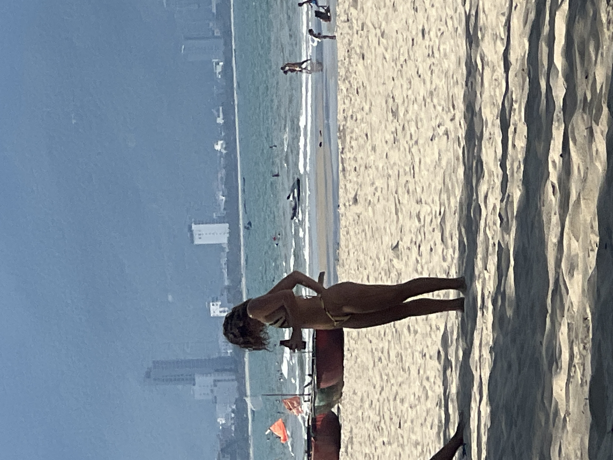 A young woman in a bikini stands on a sandy beach, holding a bottle. In the background, the ocean stretches to a city skyline with tall buildings, and the sky is hazy. Several traditional Vietnamese fishing boats are visible on the sand in the mid-ground. Other beachgoers can be seen in the distance, some in the water and others on the shore. The scene suggests a coastal urban environment in Đà Nẵng, Vietnam, likely during daylight hours given the bright lighting.