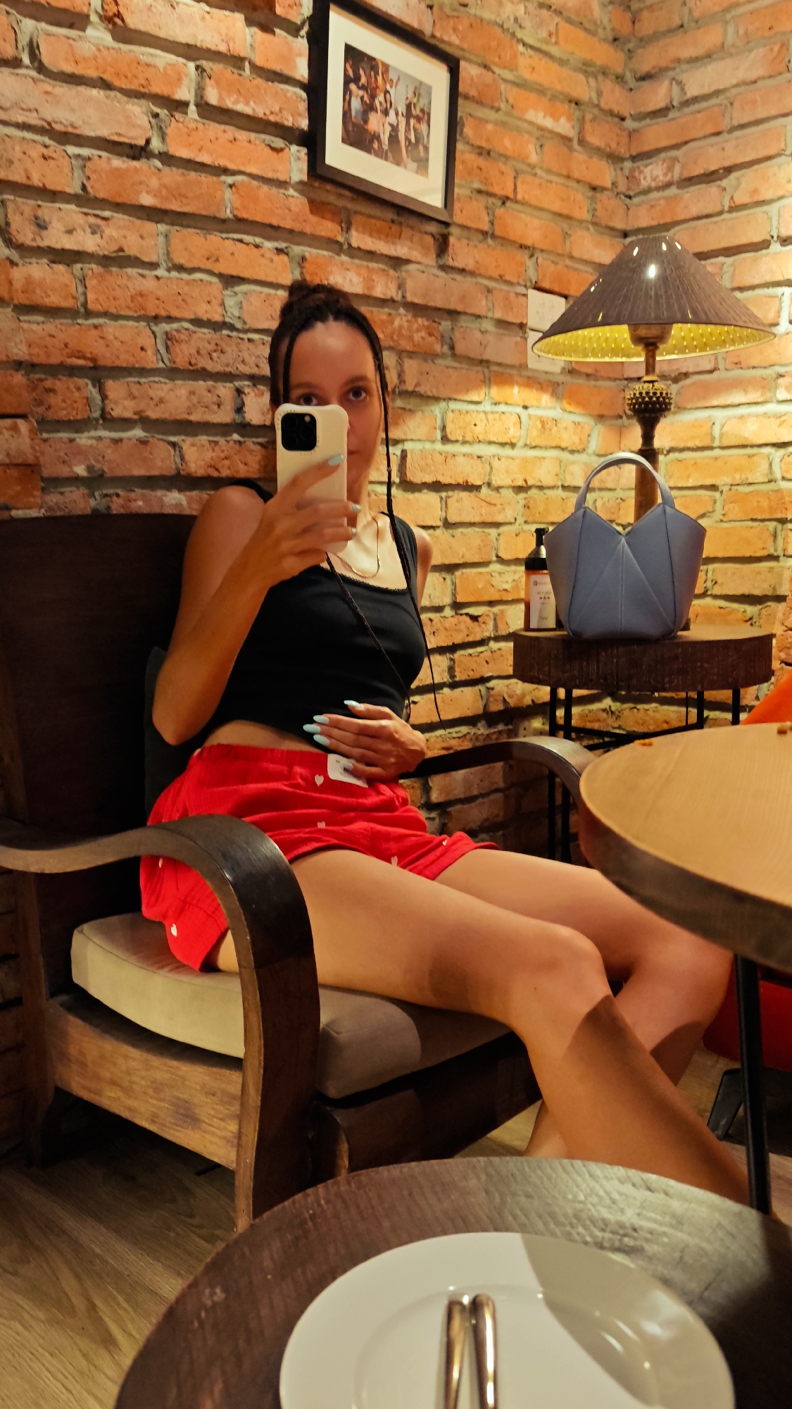 A young woman with braided hair is taking a selfie in a dimly lit cafe with exposed brick walls. She is wearing a black tank top and red shorts with white heart patterns. She is sitting in a wooden chair, with her legs crossed. To her right, on a small side table, is a blue handbag, a bottle of what appears to be hand sanitizer, and a lamp with a patterned shade. A framed picture hangs on the brick wall behind her. In the foreground, a round wooden table holds a white plate with two gold utensils. The floor is made of wooden planks. The image captures a relaxed and intimate atmosphere within a cafe in Hà Nội, Vietnam.