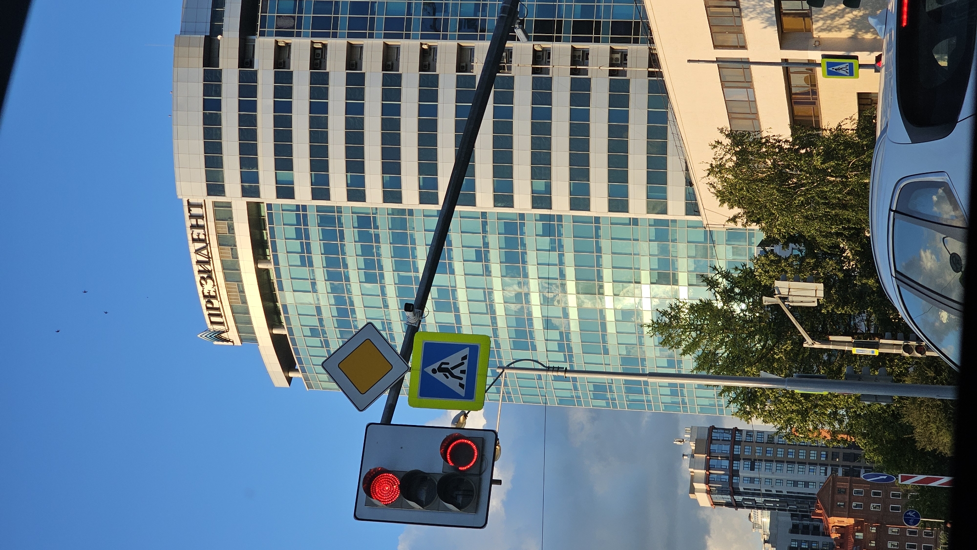This media file captures a street scene in Yekaterinburg, Russia, with a focus on a modern glass-fronted building and traffic signals. The image is oriented vertically, showing a clear blue sky with a few distant birds, suggesting daytime. The building in the background has a prominent sign with Cyrillic text that reads "ПРЕЗИДЕНТ" (PRESIDENT).
In the foreground, a traffic light shows red for both directions of travel, indicating that traffic is stopped. Beside the traffic light are two road signs: a diamond-shaped yellow sign with a black border, which is a warning sign, and a blue square sign with a white pictogram of a person walking, indicating a pedestrian crossing. A silver car is partially visible on the right side of the frame, reflecting the sky and trees.
The scene is urban, with multiple buildings visible, including a more distant residential building with a red brick facade. Lush green trees add a touch of nature to the cityscape. The lighting suggests it is either morning or late afternoon, with the sun casting shadows and highlighting the architectural details of the buildings. The overall mood is that of a typical urban environment during daylight hours.