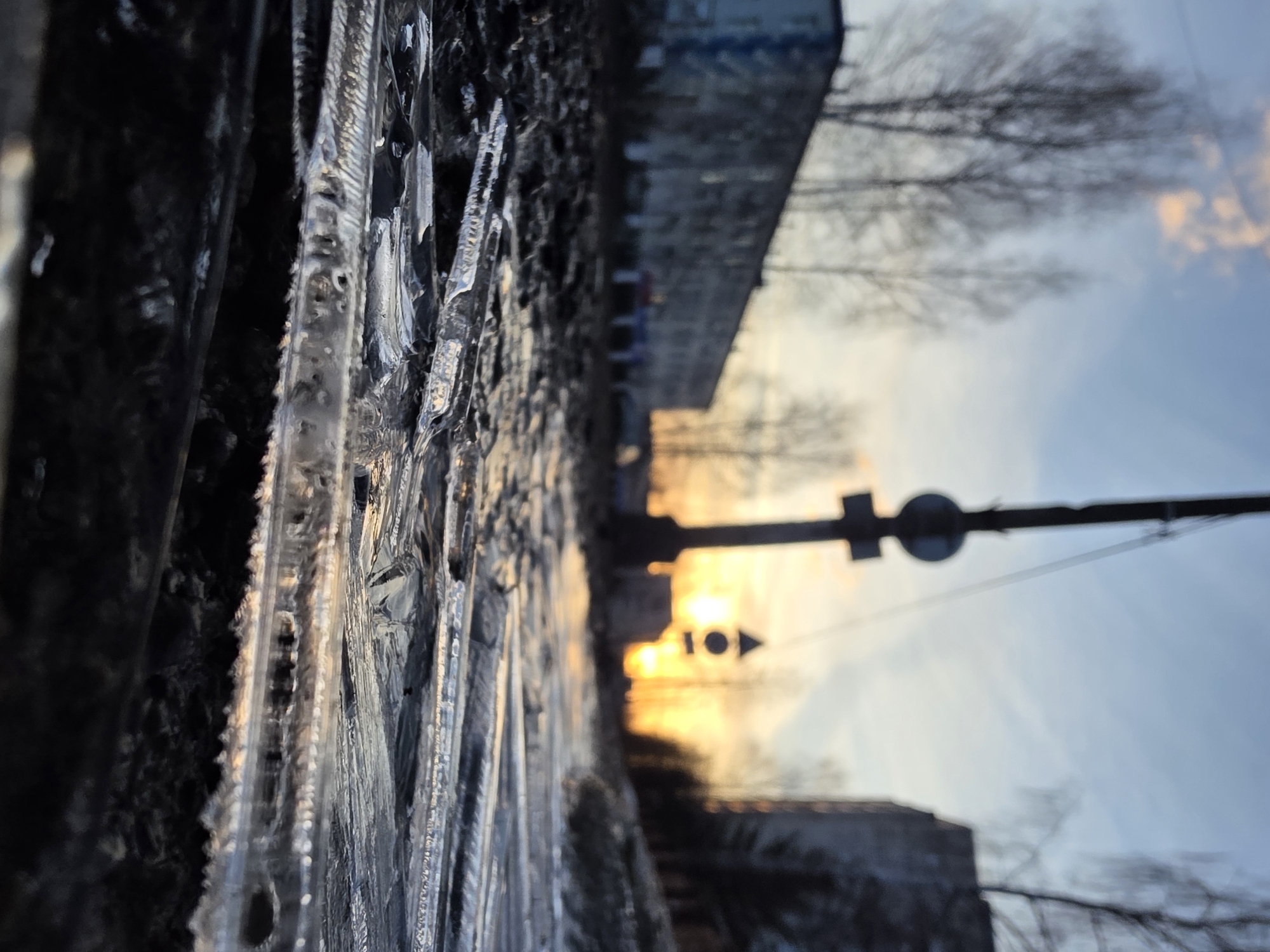 A low-angle shot looking out from a ledge covered in icicles, likely during a winter sunset in Chaikovsky, Russia. In the background, a building stands to the left, its details slightly obscured. Between the building and the icicles, the setting sun casts a bright glow. A traffic light is prominently silhouetted against the sky. The sky itself fades from a yellowish-orange near the horizon to a pale blue higher up. Bare tree branches are silhouetted against the sky.