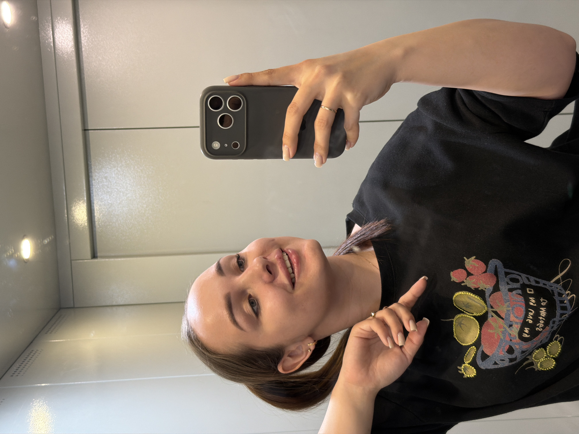 A young woman is taking a selfie with her phone. She has fair skin, brown hair pulled back into a low ponytail, and is wearing a black t-shirt with a colorful design on it. She is holding a black smartphone with a triple-lens camera. The background appears to be the interior of a small elevator with white, metallic walls. Given the mention of the city of Chaikovsky, Russia, it can be assumed the image was taken there.