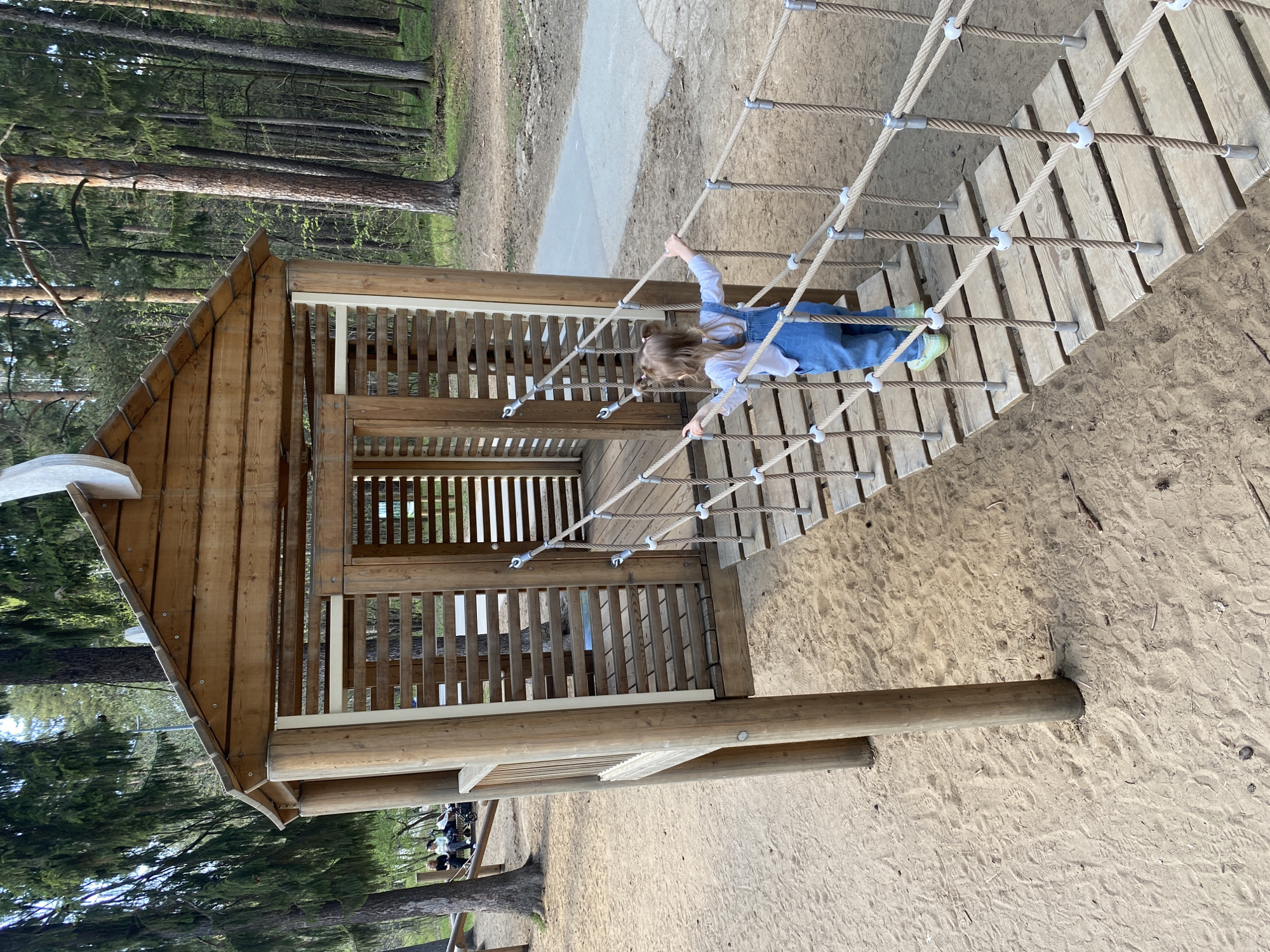 A young girl is navigating a rope bridge connecting two wooden play structures in a playground. The structures feature slatted walls and roofs. The area around the playground is covered in sand, with a forest visible in the background. While the exact location within the city of Perm, Russia, cannot be determined with certainty, it is likely within a park or recreational area.