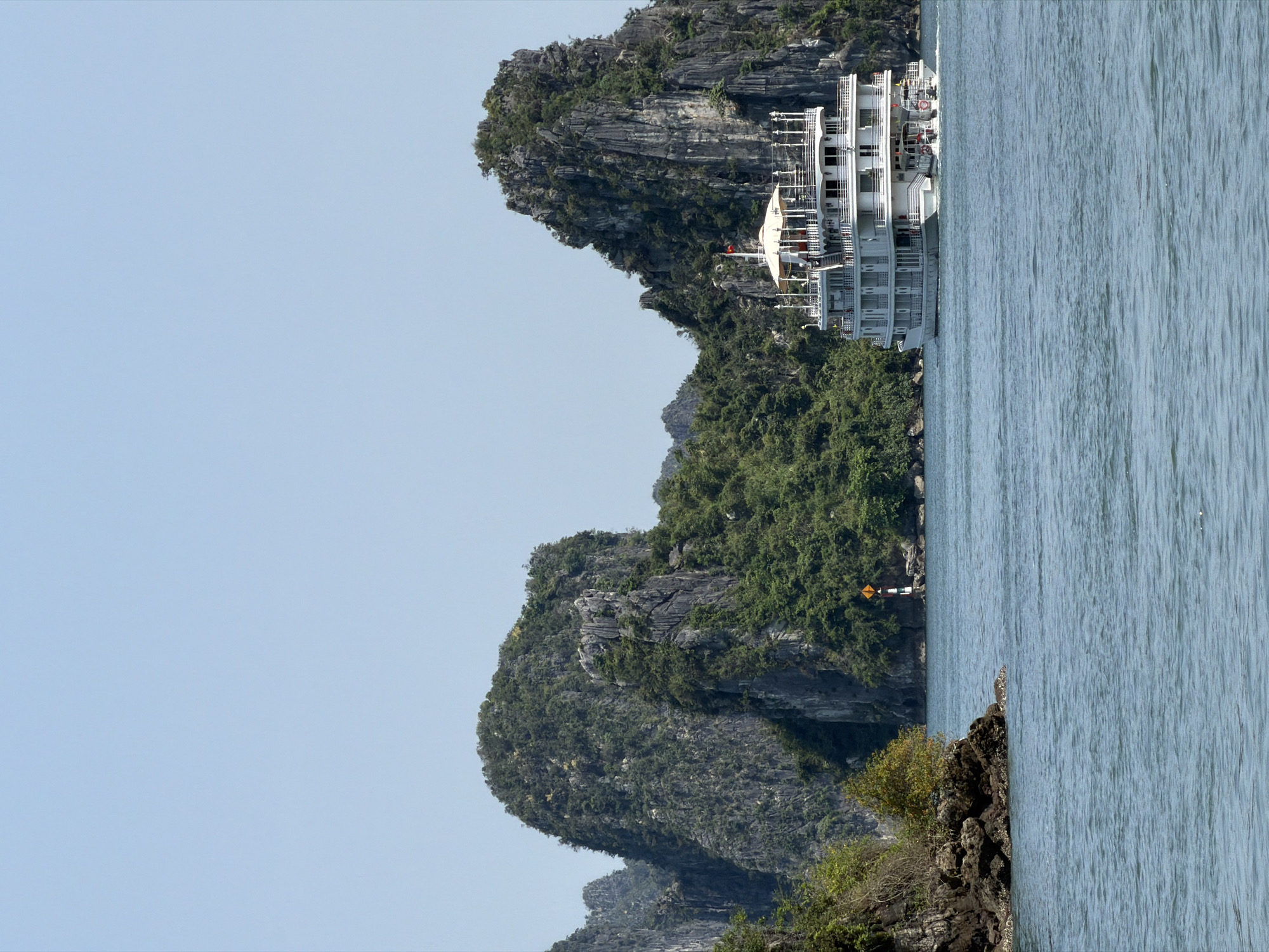 This image captures a picturesque scene of a large cruise ship navigating or anchored within a bay surrounded by dramatic limestone karsts. The main subject is a multi-decked white cruise ship, visible in the mid-ground, nestled between the towering rock formations. The ship features numerous windows, balconies, and railings, and a red flag with a yellow star, the national flag of Vietnam, is visible near its upper deck.
The bay is characterized by its distinctive karst islands, which rise steeply from the calm, blue-green water. These islands are heavily vegetated with dense green trees and shrubs, contrasting with the grey, rugged rock faces. In the mid-ground, on one of the smaller islands, a yellow diamond-shaped warning sign with a black border and a black exclamation mark is clearly visible, likely indicating a hazard or caution area for navigation.
The water surface is relatively calm, showing subtle ripples, reflecting the clear, pale blue sky that dominates the upper portion of the image. The lighting suggests a bright, clear day, likely late morning or early afternoon, with no harsh shadows. The overall setting evokes a sense of tranquility and natural grandeur, typical of the iconic landscapes found in areas such as Ha Long Bay in Vietnam. The scene highlights the interplay between natural beauty and human presence, with the cruise ship offering a way to experience the stunning environment.